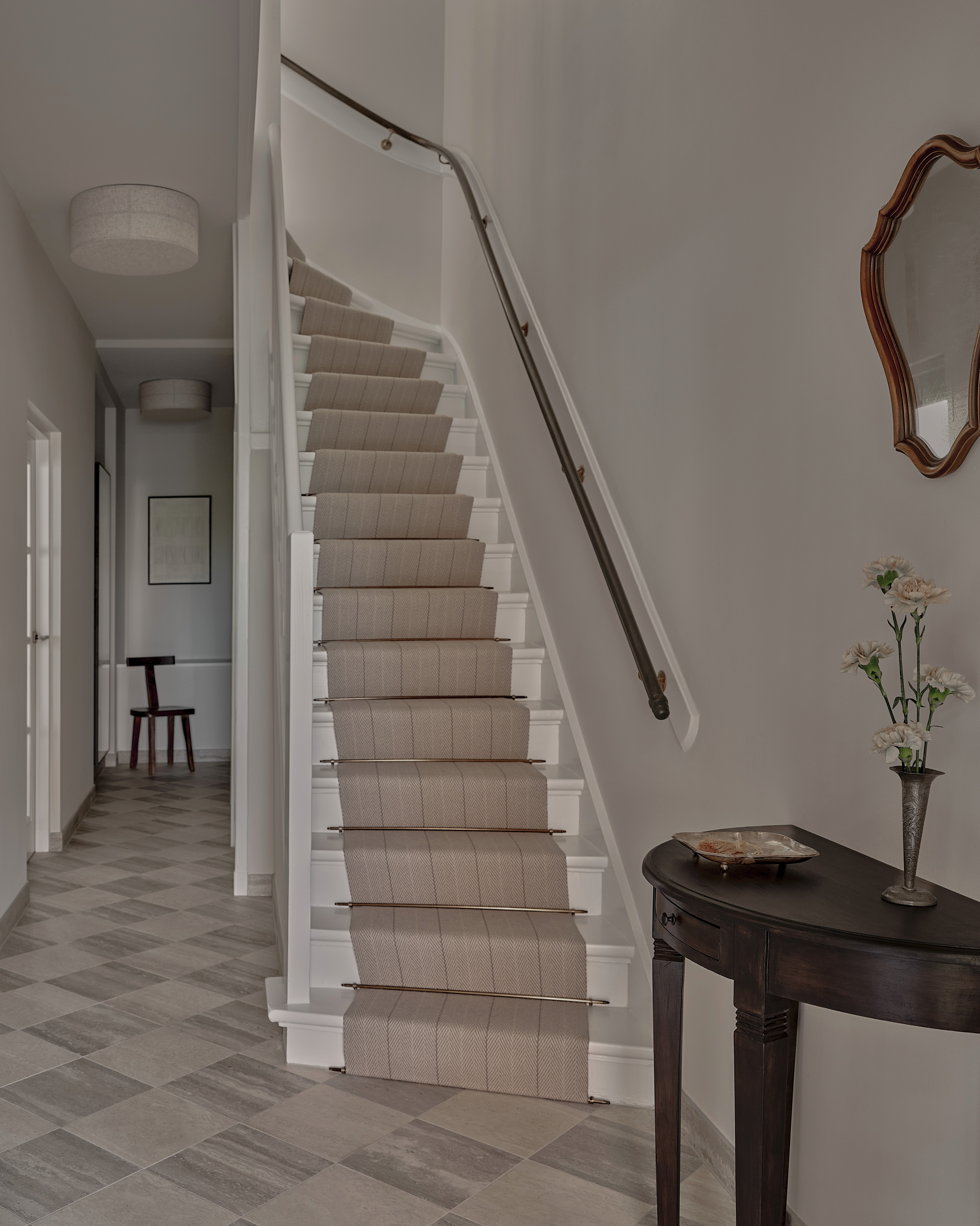 Entryway with white staircase, neutral wool runner with brass stair rods, antique demilune console, scalloped wood mirror, and stone tile floor