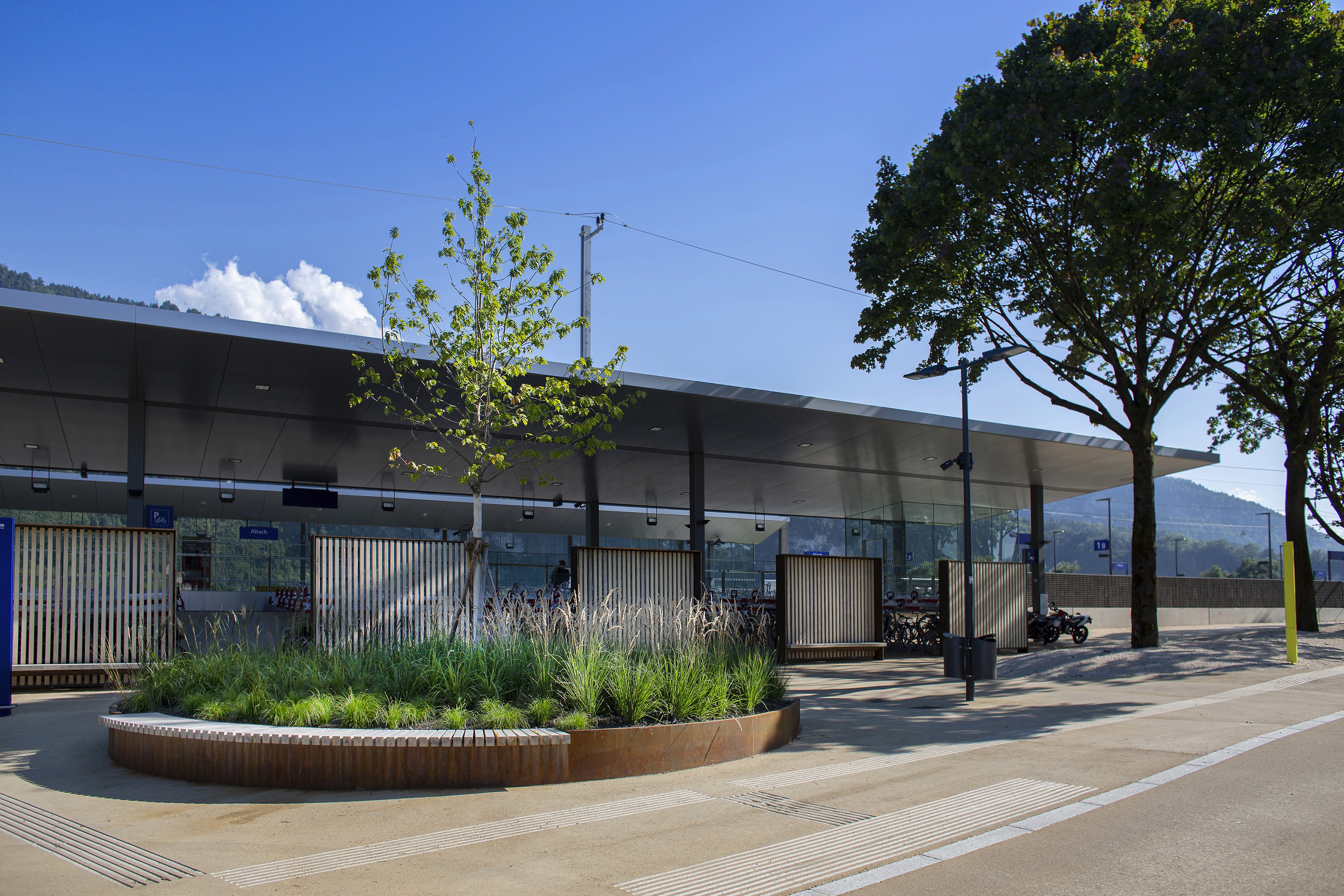 Moderner Bahnhof Altach mit lang gestrecktem, flachen Dach, Glasfassade und begrünten Rundbeeten am Vorplatz bei sonnigem Himmel.