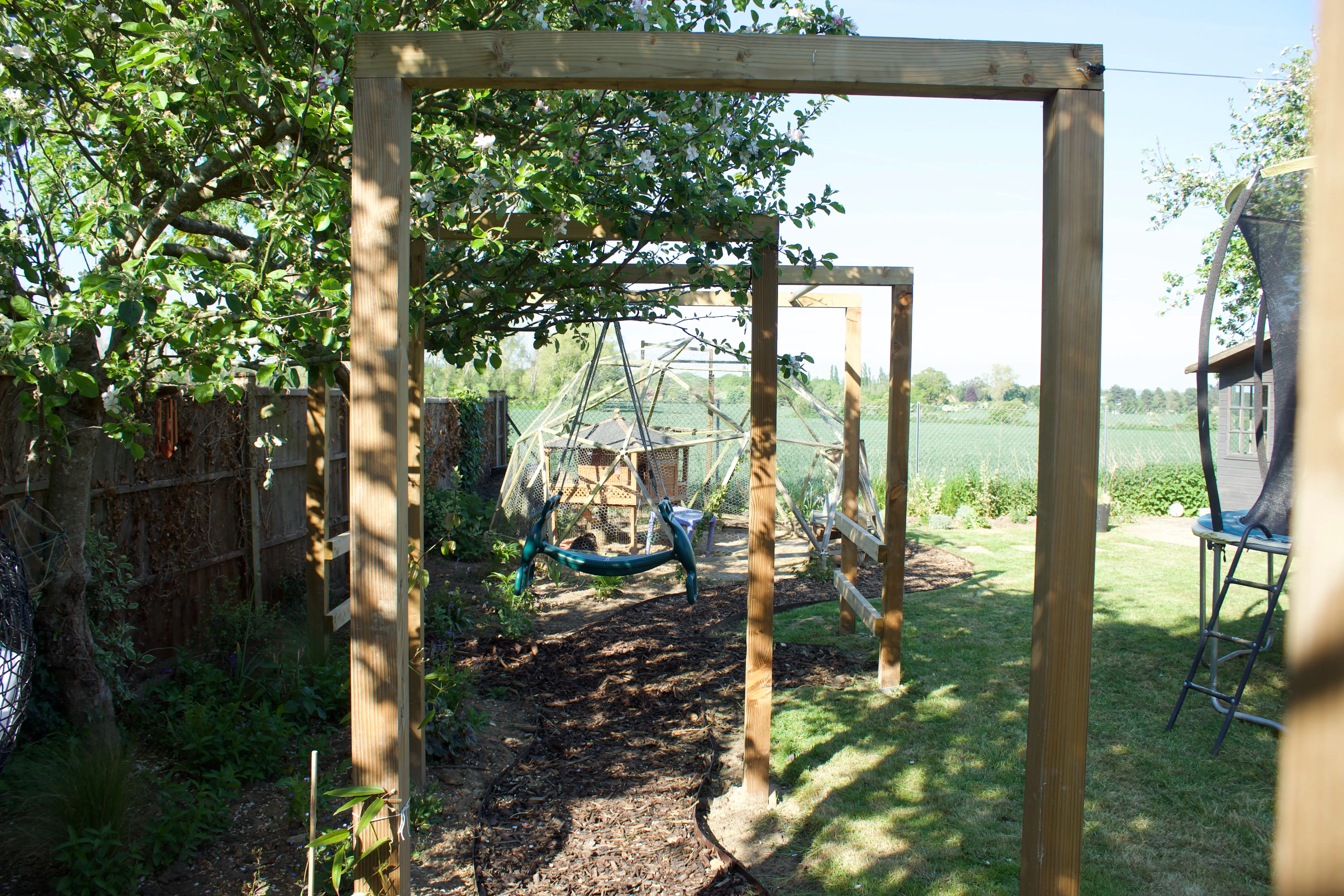 A wooden garden archway leads into a lush green outdoor space, framed by trees and bright sky.