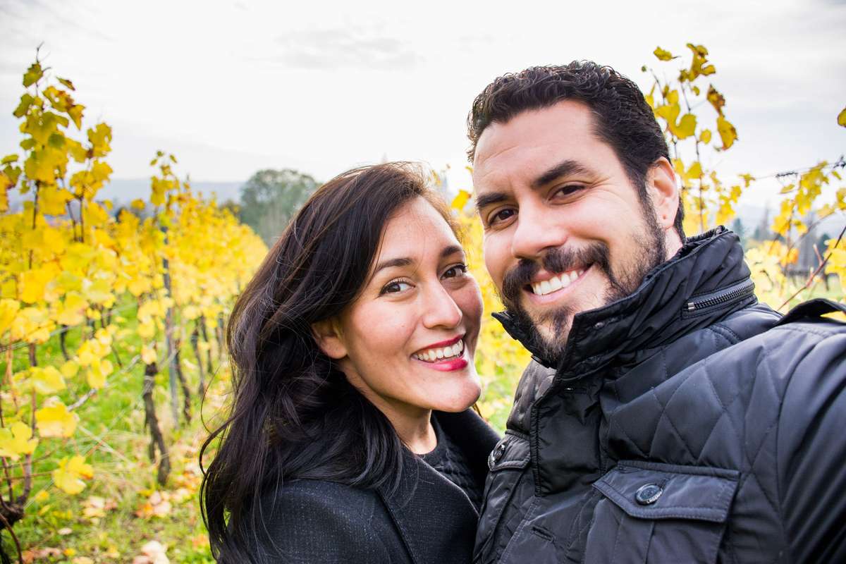 A smiling man and woman pose in front of a vineyard with yellow flowers under an overcast sky.