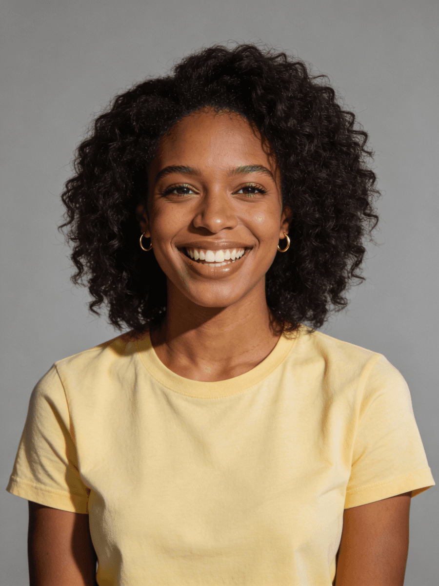 Portrait of a smiling woman with natural curly hair wearing a pale yellow T-shirt and gold hoop earrings against a gray background.