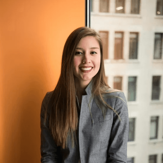 Smiling woman with long brown hair wearing a grey blazer, standing between a bright orange wall and a city window.