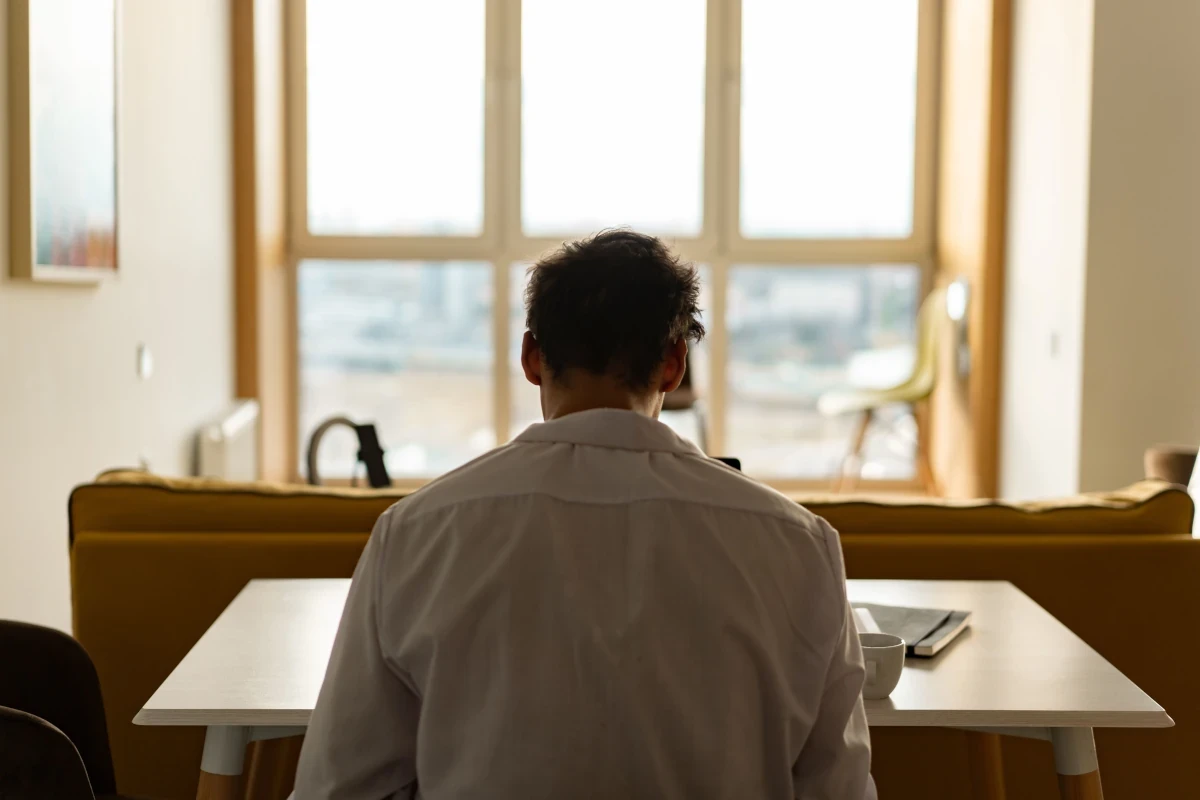 Man sitting alone with his back to the camera in a bright room, appearing quiet and withdrawn.