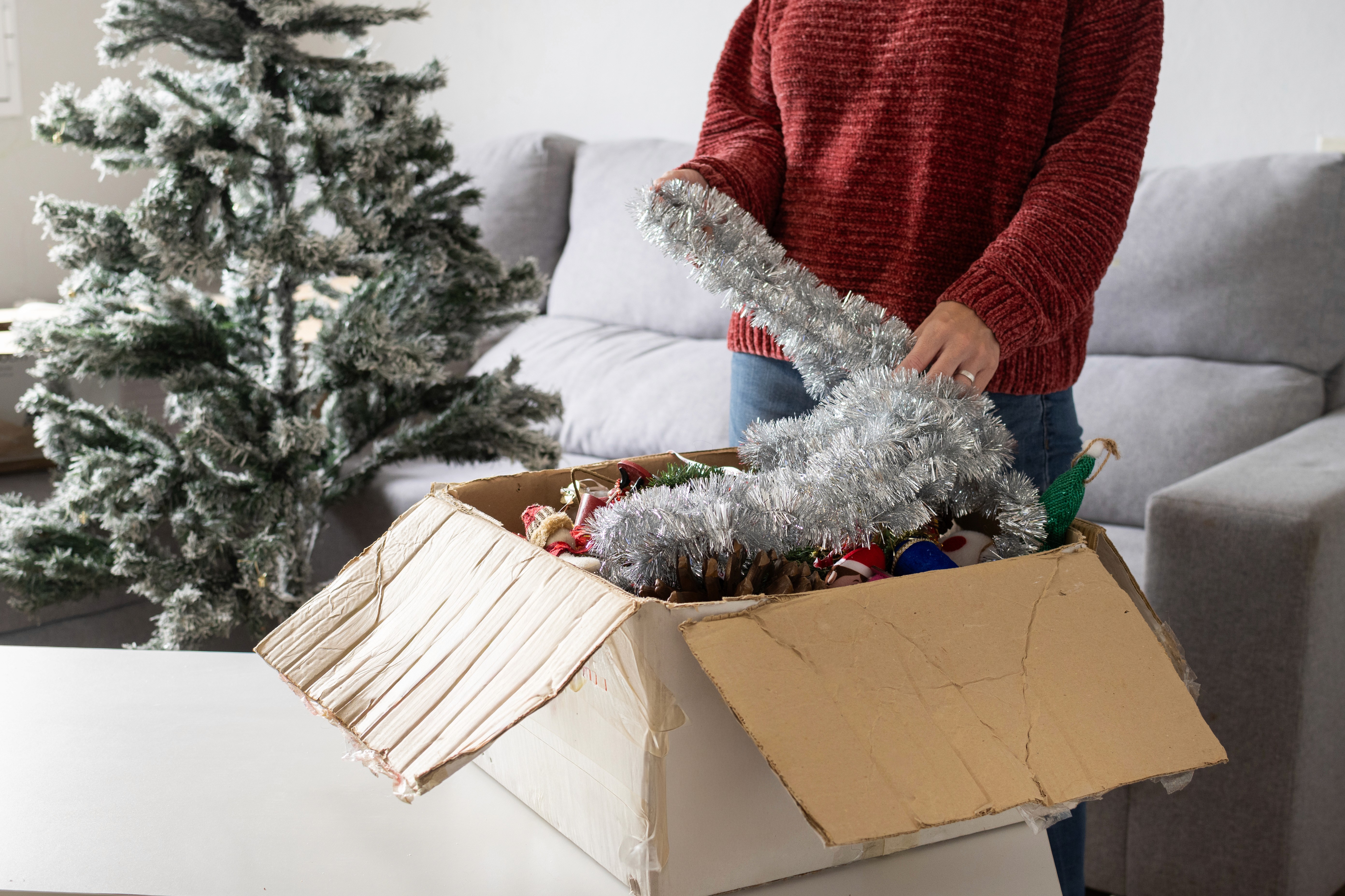 A person in a red sweater packs away silver tinsel from a cardboard box filled with Christmas decorations, with a frosted artificial tree nearby.