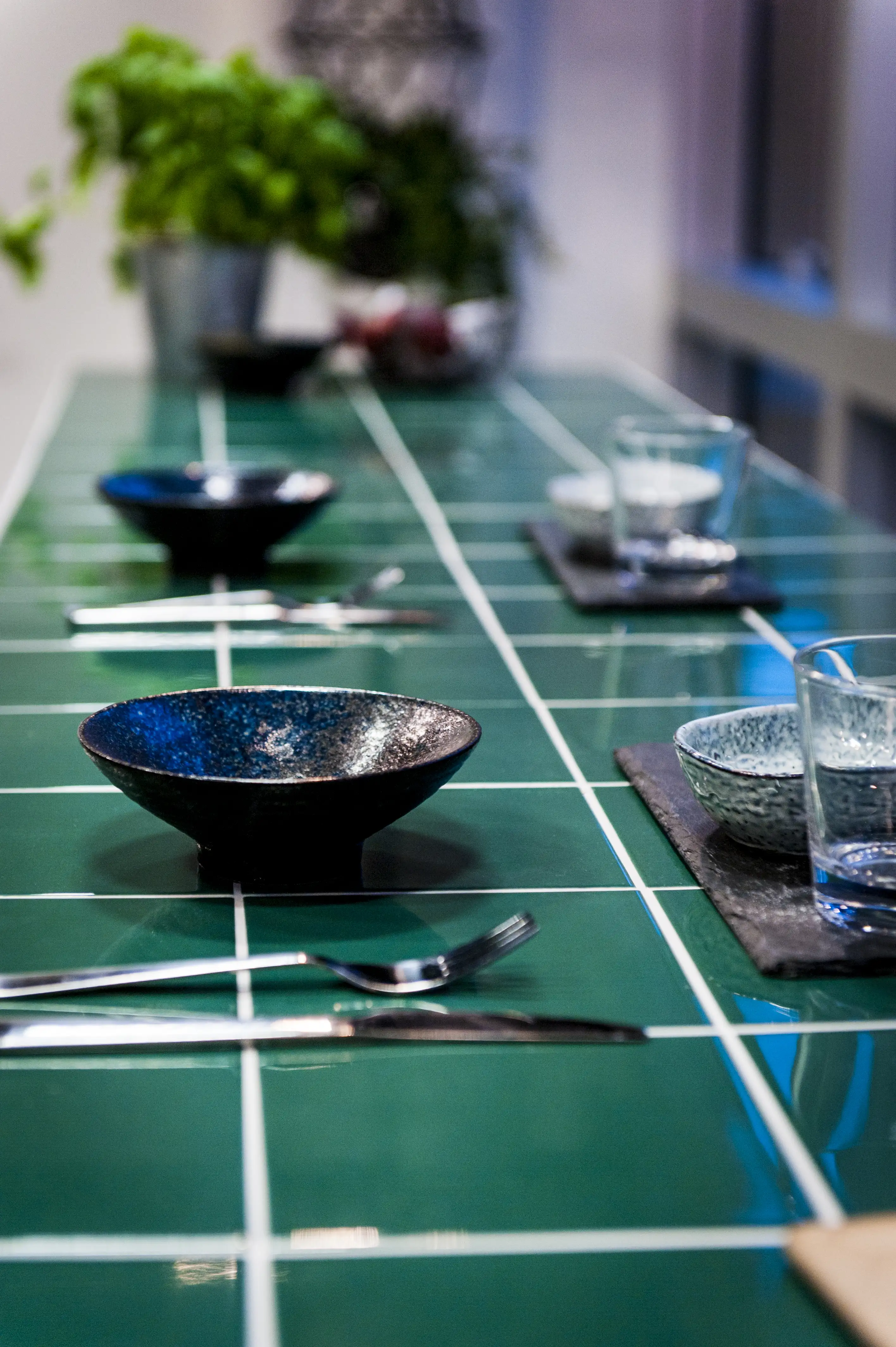 Detail of the green-tiled table top at KANTINE KOMTUR set with black ceramic bowls, cutlery, glasses and slate placemats.