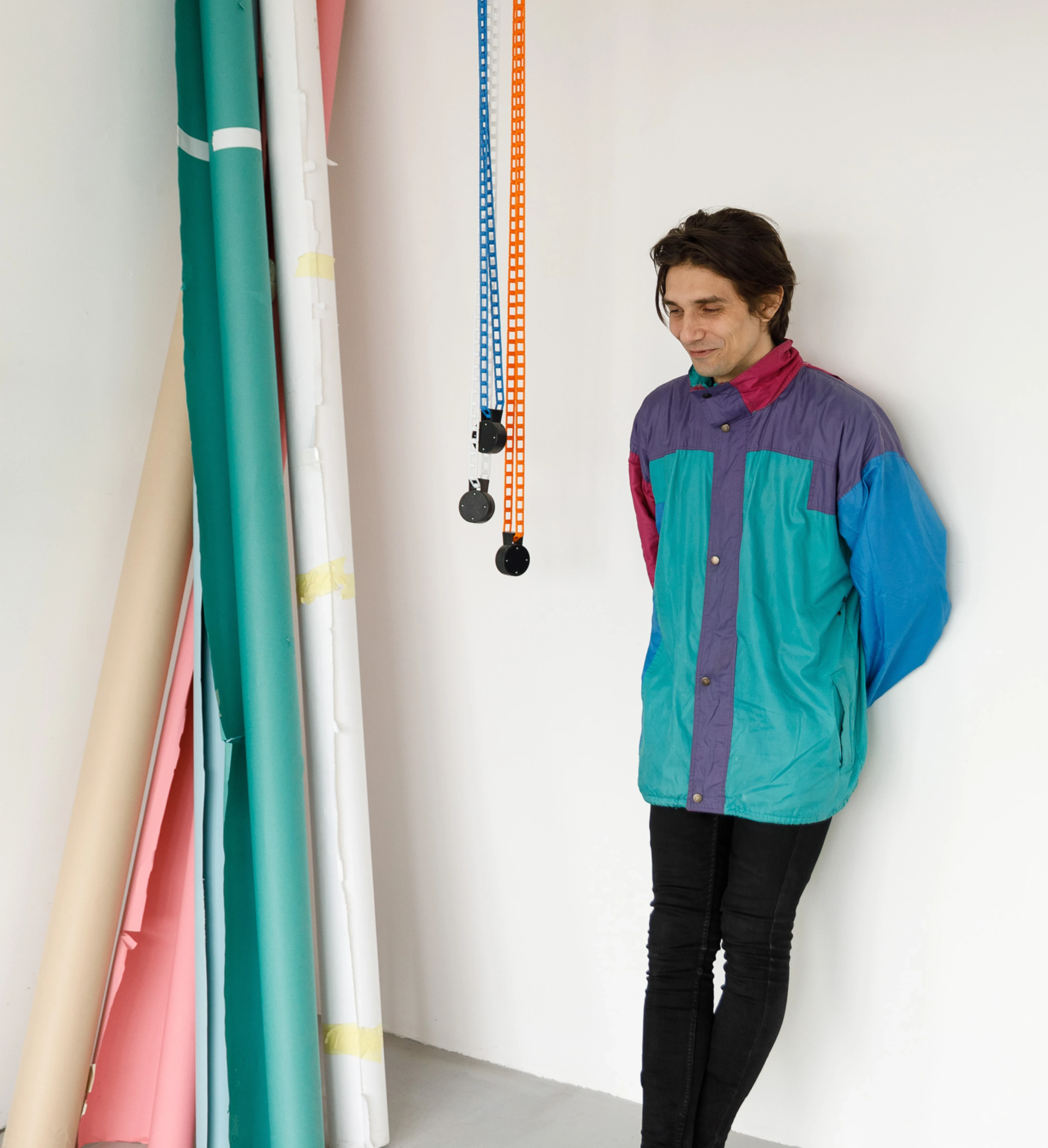 A person in a colorful retro windbreaker and black jeans leans against a white wall, smiling softly, surrounded by hanging pulleys and colorful paper rolls.