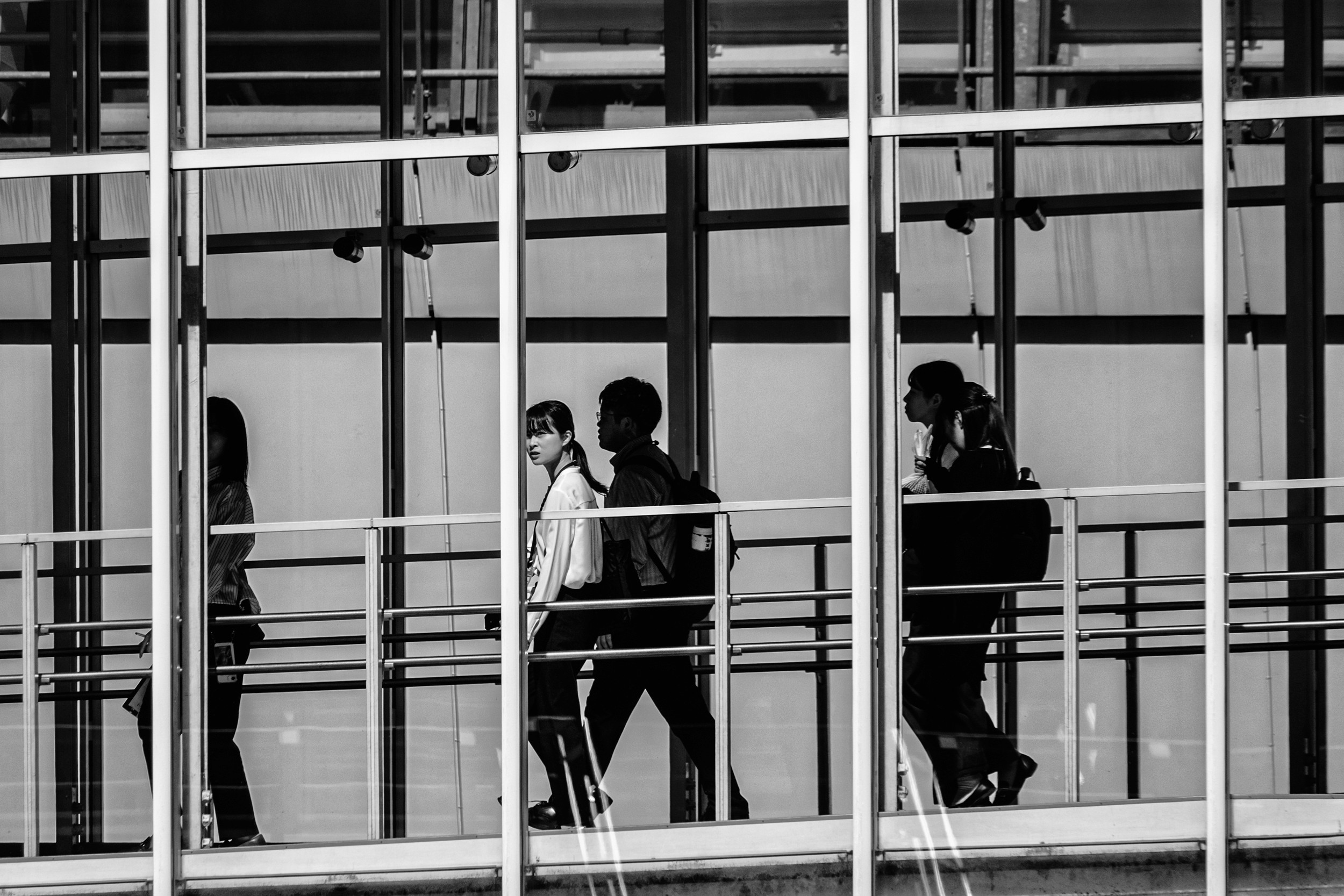 Black and white photograph of office workers crossing a glass skywalk between buildings.