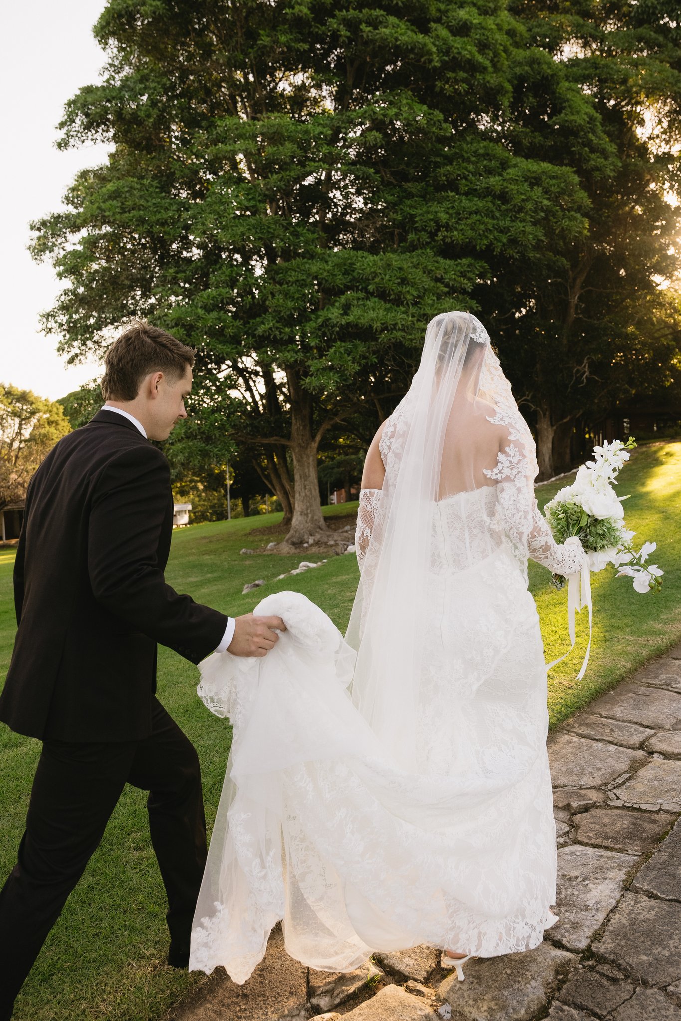 Groom carrying the bride's train as the couple walk through late-afternoon light