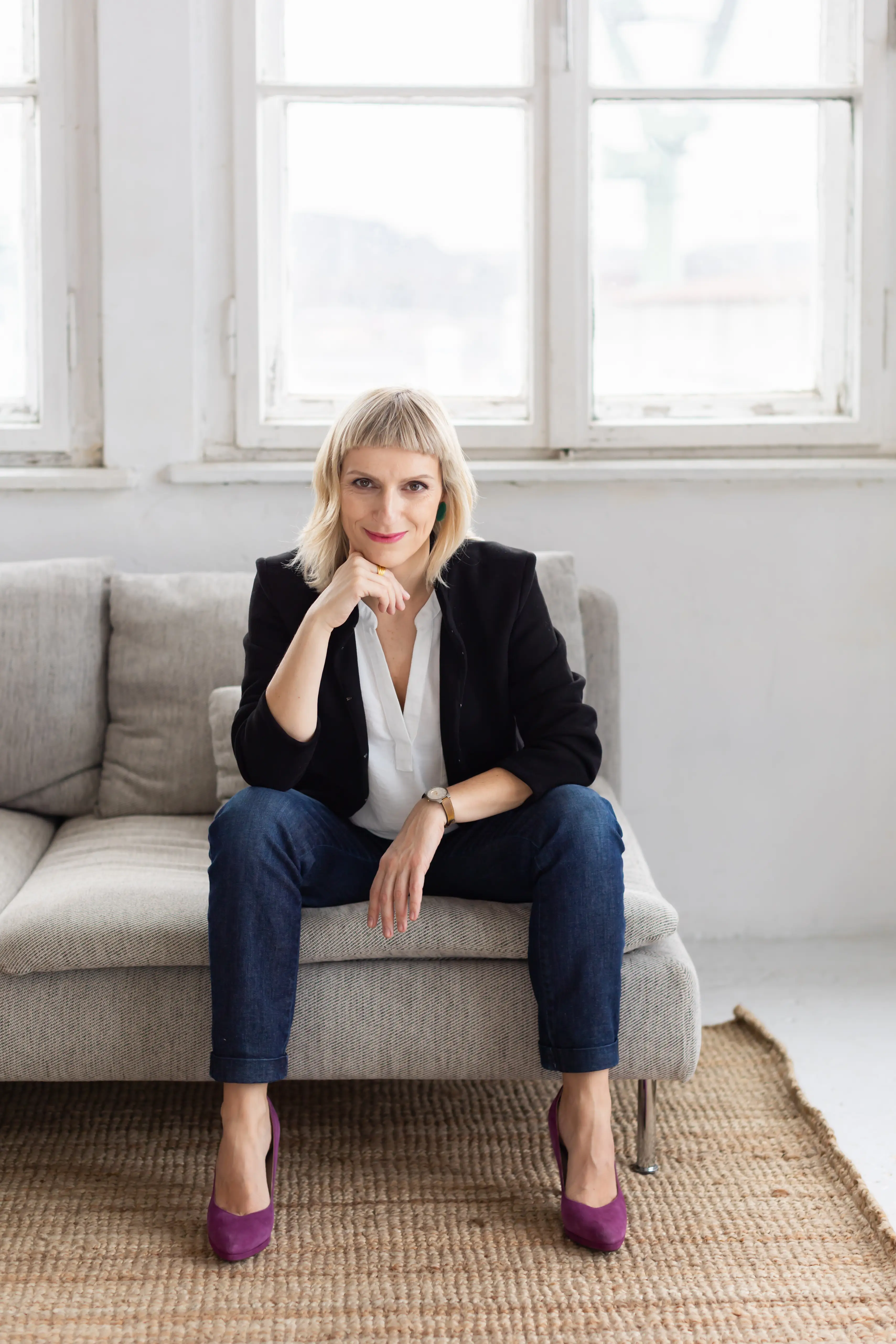 Kobieta o krótkich blond włosach siedzi na jasnoszarej sofie, ubrana w czarną marynarkę, biały top, niebieskie dżinsy i fioletowe buty. Uśmiecha się, opierając podbródek na dłoni, w jasnym pokoju z dużymi oknami.