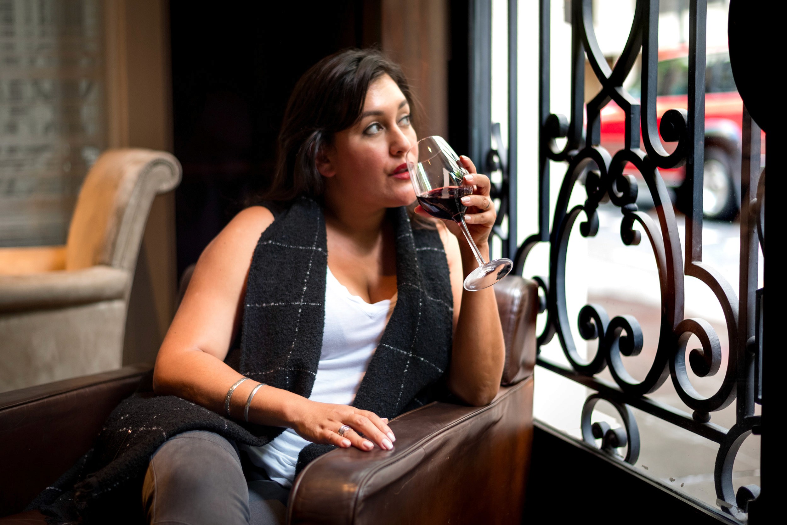 A woman in a grey vest and white top sits in a leather chair by a window, holding a glass of red wine.