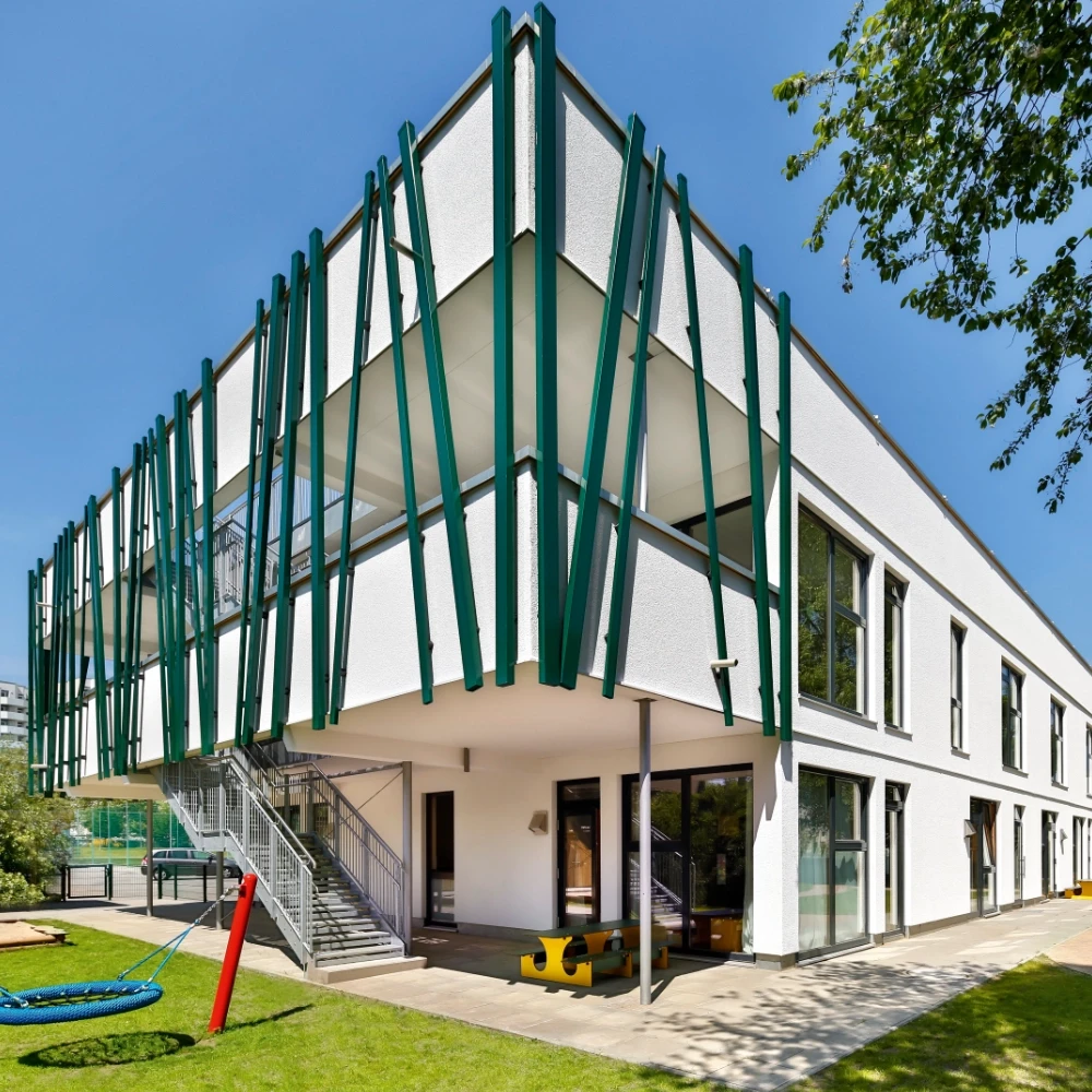 Modernes Gebäude mit weißer Fassade, grünen vertikalen Stützen, Außentreppen und Spielplatz an einem sonnigen Tag.