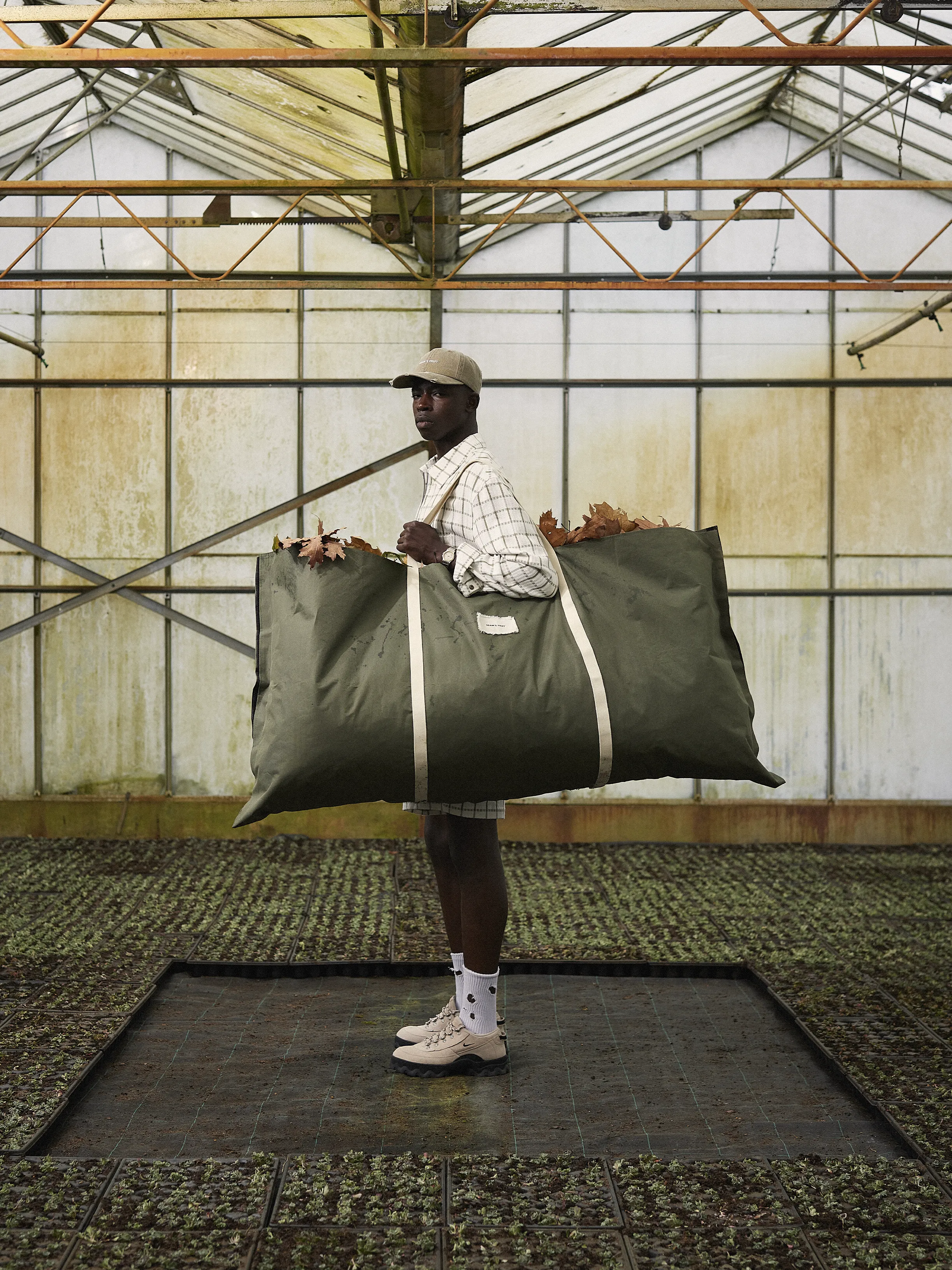 Dans une serre, avec un sac plein de feuilles mortes, le mannequin porte un look de la collection FW25 de Brams Fruit