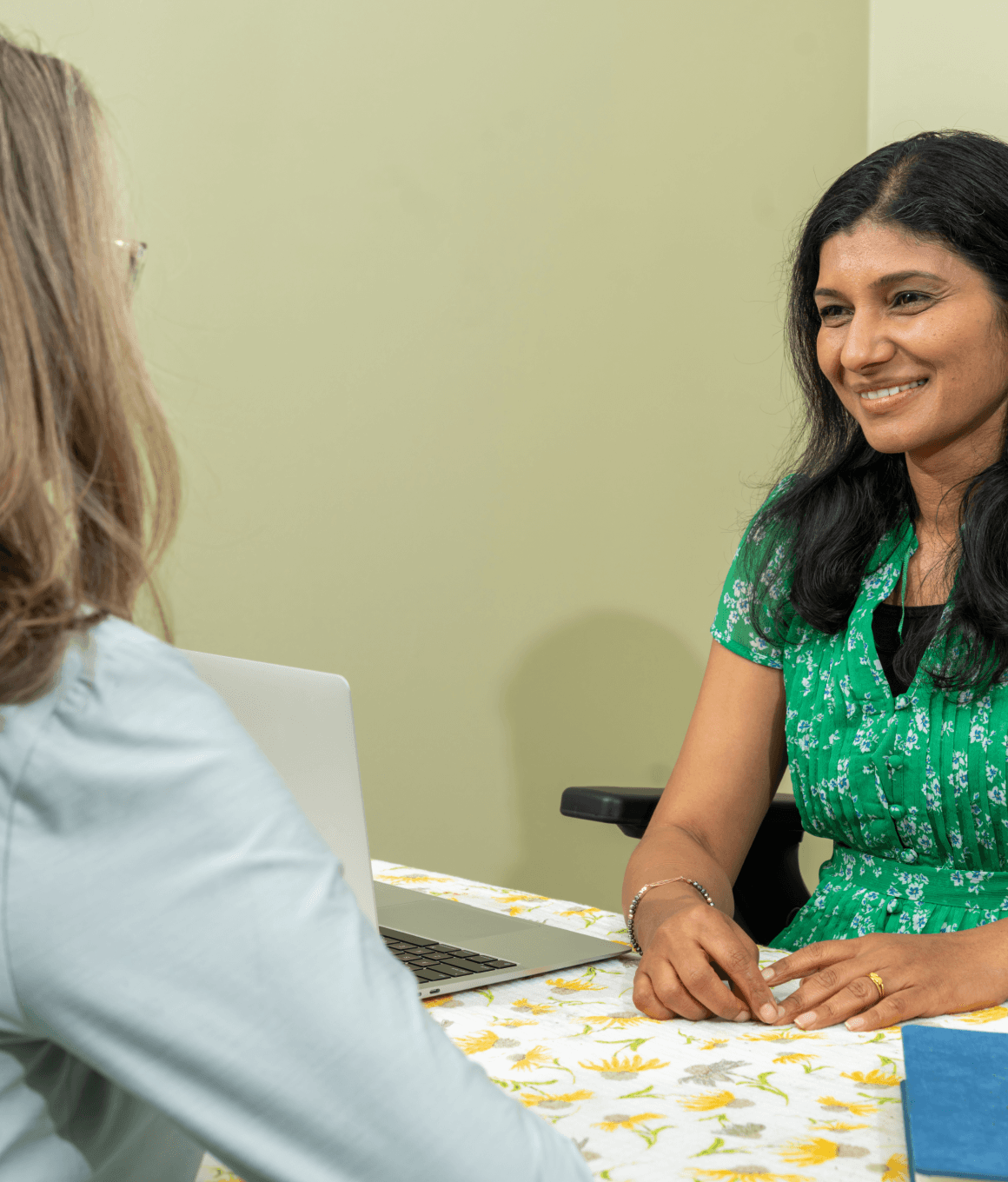 A woman in a green dress is sitting at a table in a consultation setting, engaged in conversation with another person, emphasizing the concept of Ayurvedic consultations.
