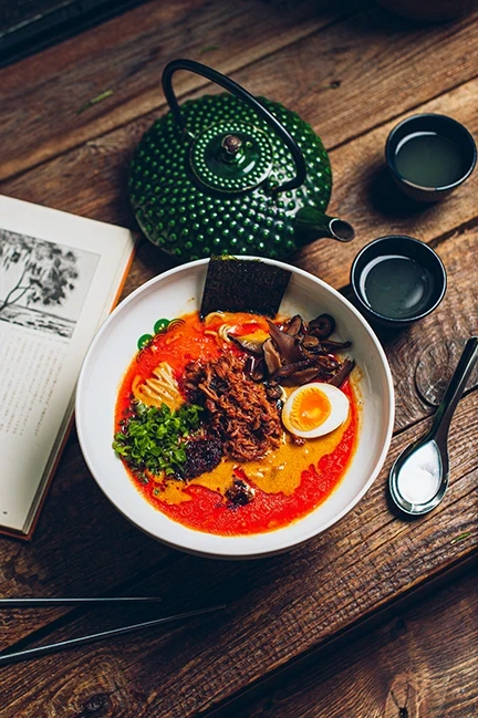 Fotografia miski z ramenem z jajkiem, grzybami i zieloną cebulką, na drewnianym stole obok herbaty i otwartej książki