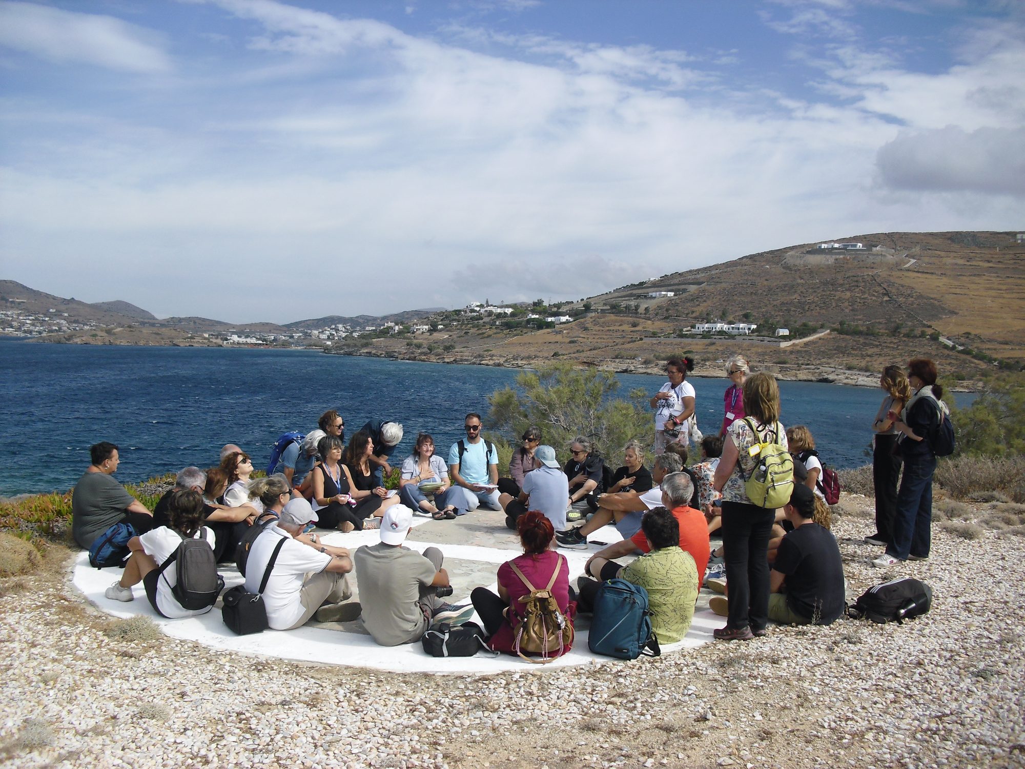 Erasmus+ participants learning in a circle on the coast of Komito, Syros, with the bay and Cycladic hills behind