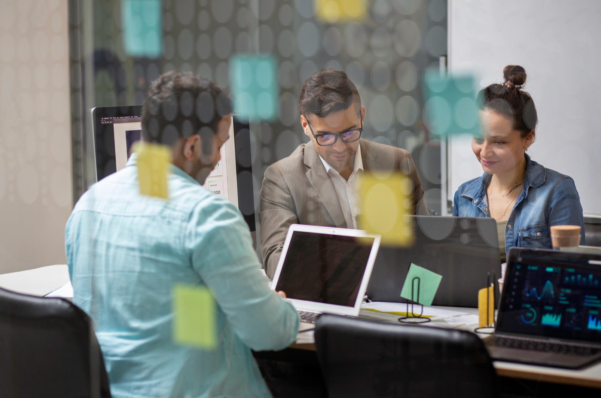 Team meeting behind a glass wall with sticky notes, reviewing laptops and planning tasks.