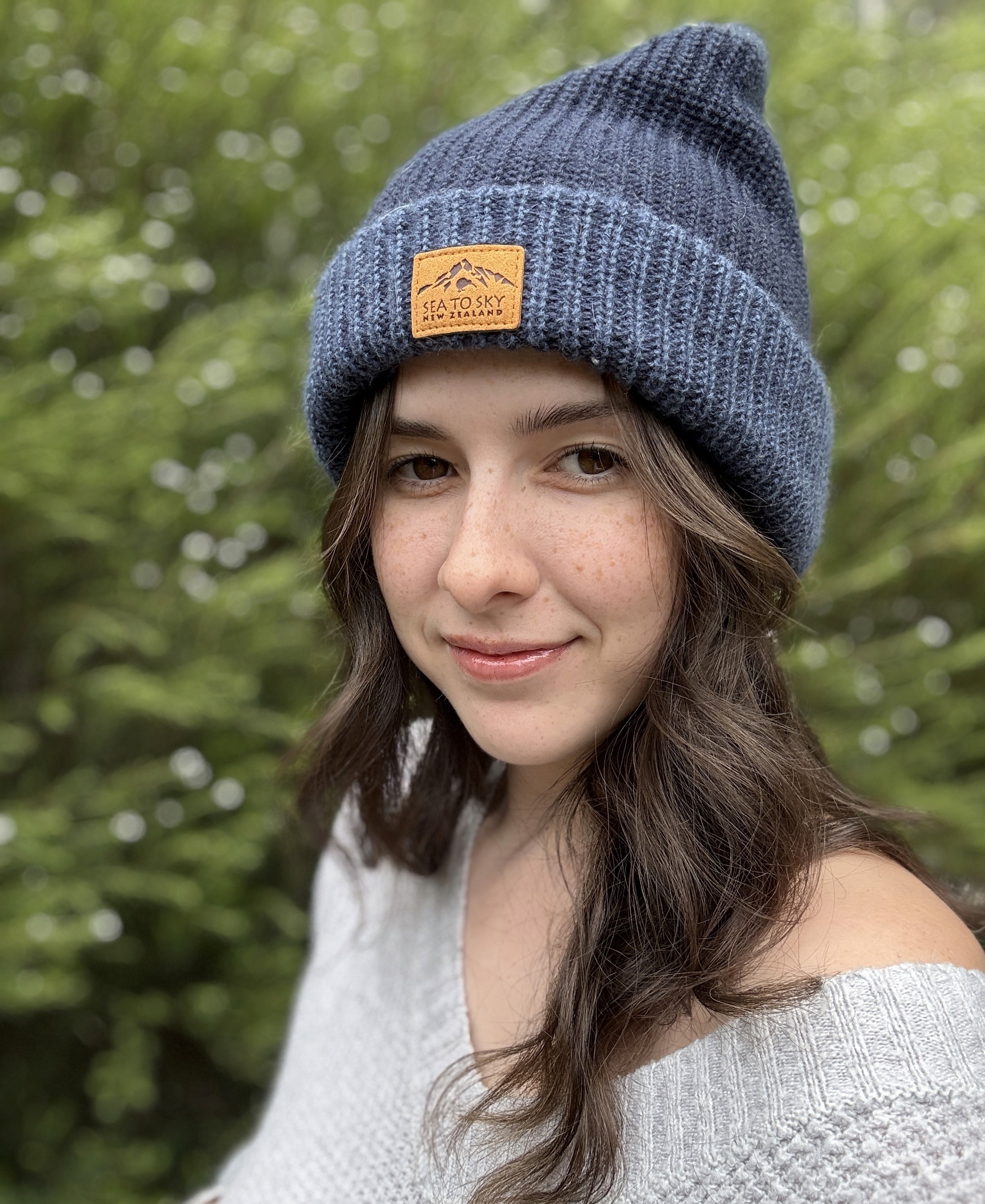 Kathryn, the designer wearing a blue beanie, a white sweater, smiling in front of a foliage background.