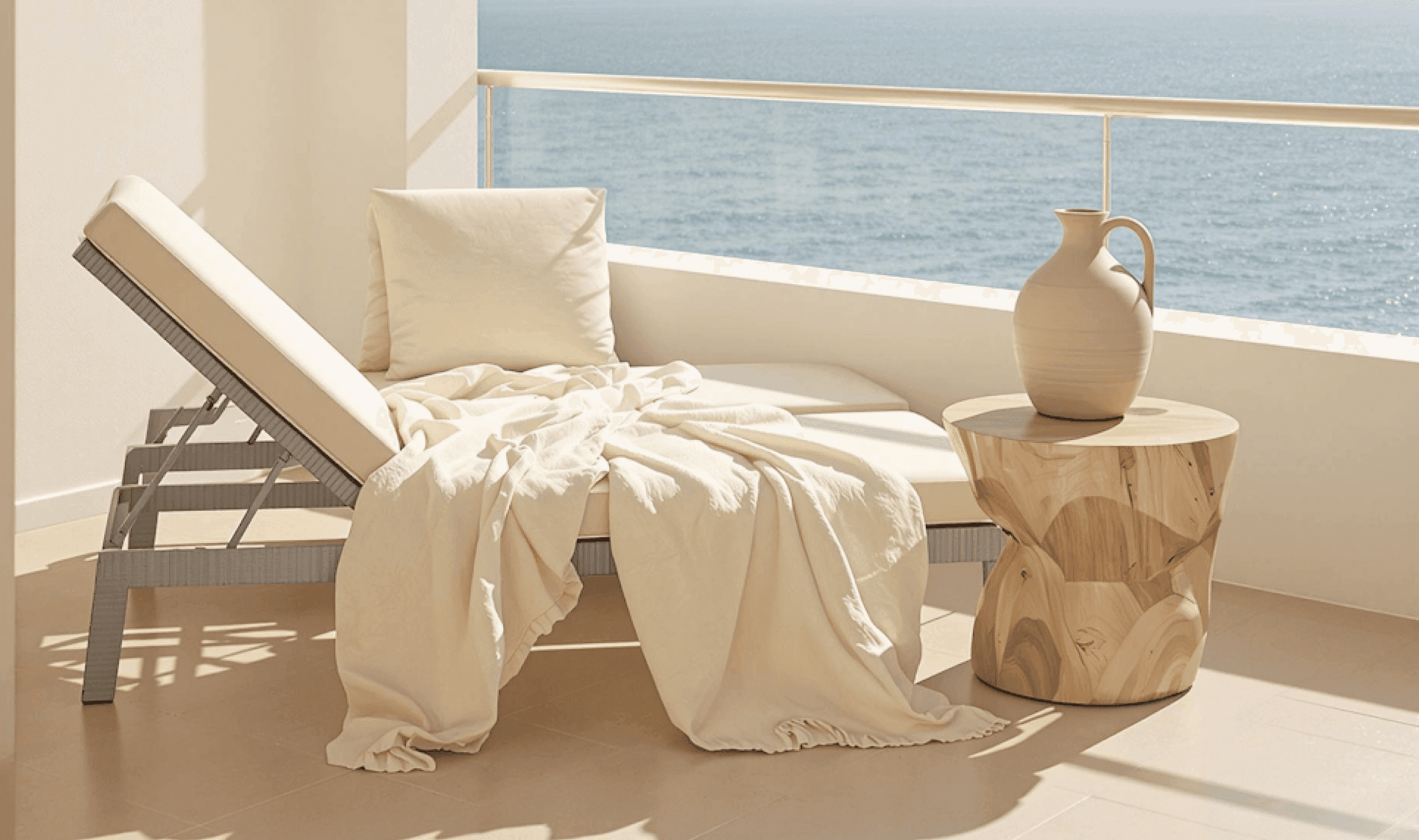 Sunlit terrace with a cushioned lounge chair, light blanket, wooden side table, and sea view.