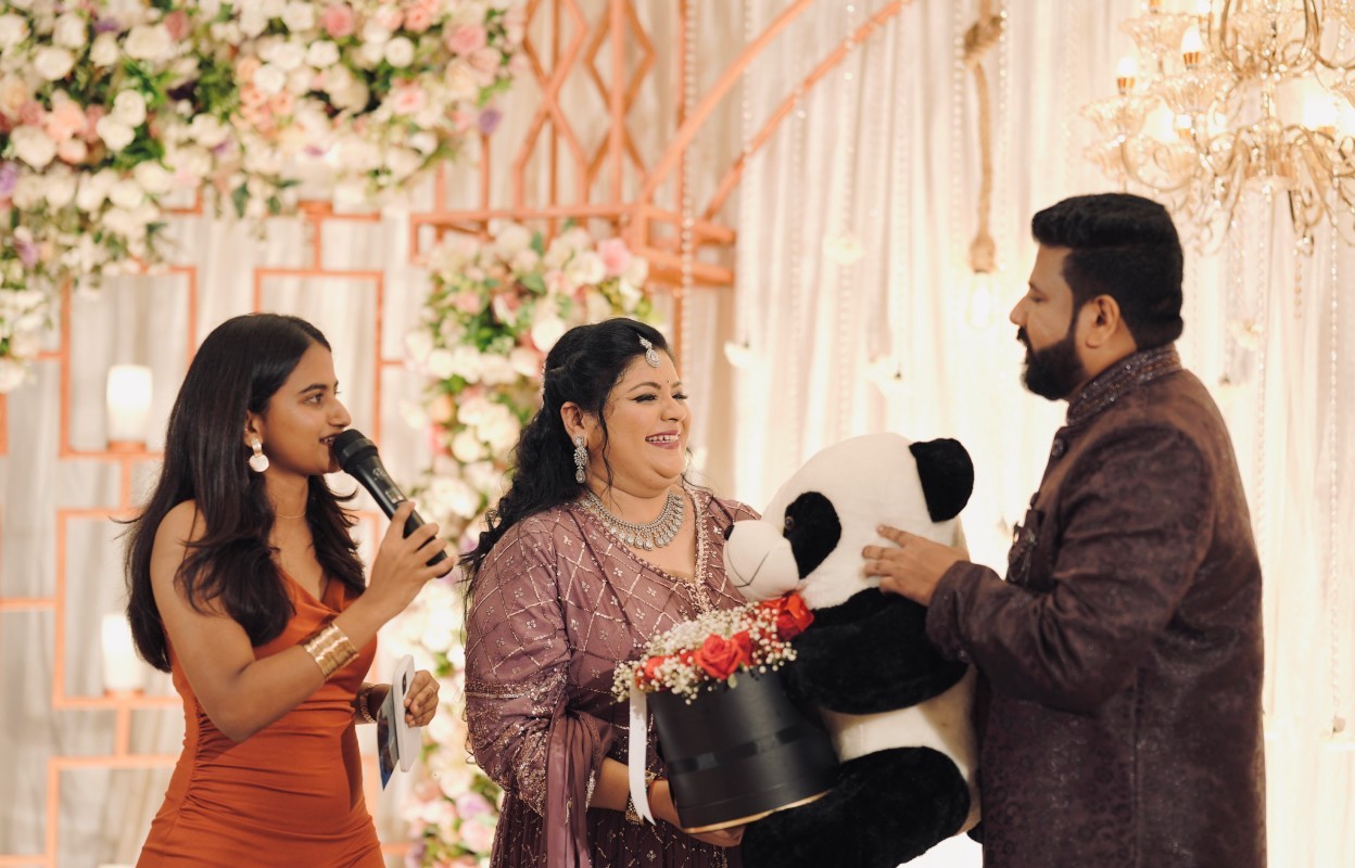 Couple exchanging a gift at an event while the emcee speaks, everyone smiling