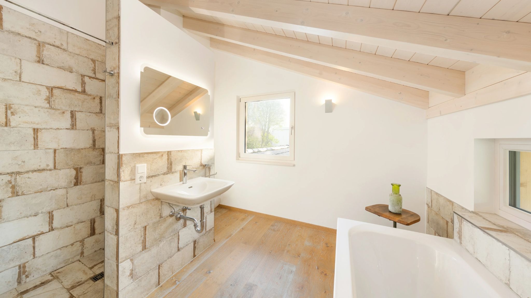 Modernes Badezimmer mit freistehender Badewanne, Dusche mit Steinoptik und Waschtisch mit beleuchtetem Spiegel unter Dachschrägen.