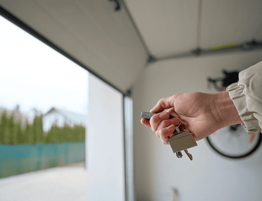 Close-up of a hand holding a garage door remote and house keys inside an open garage. Highlighting residential access control, smart garage automation.