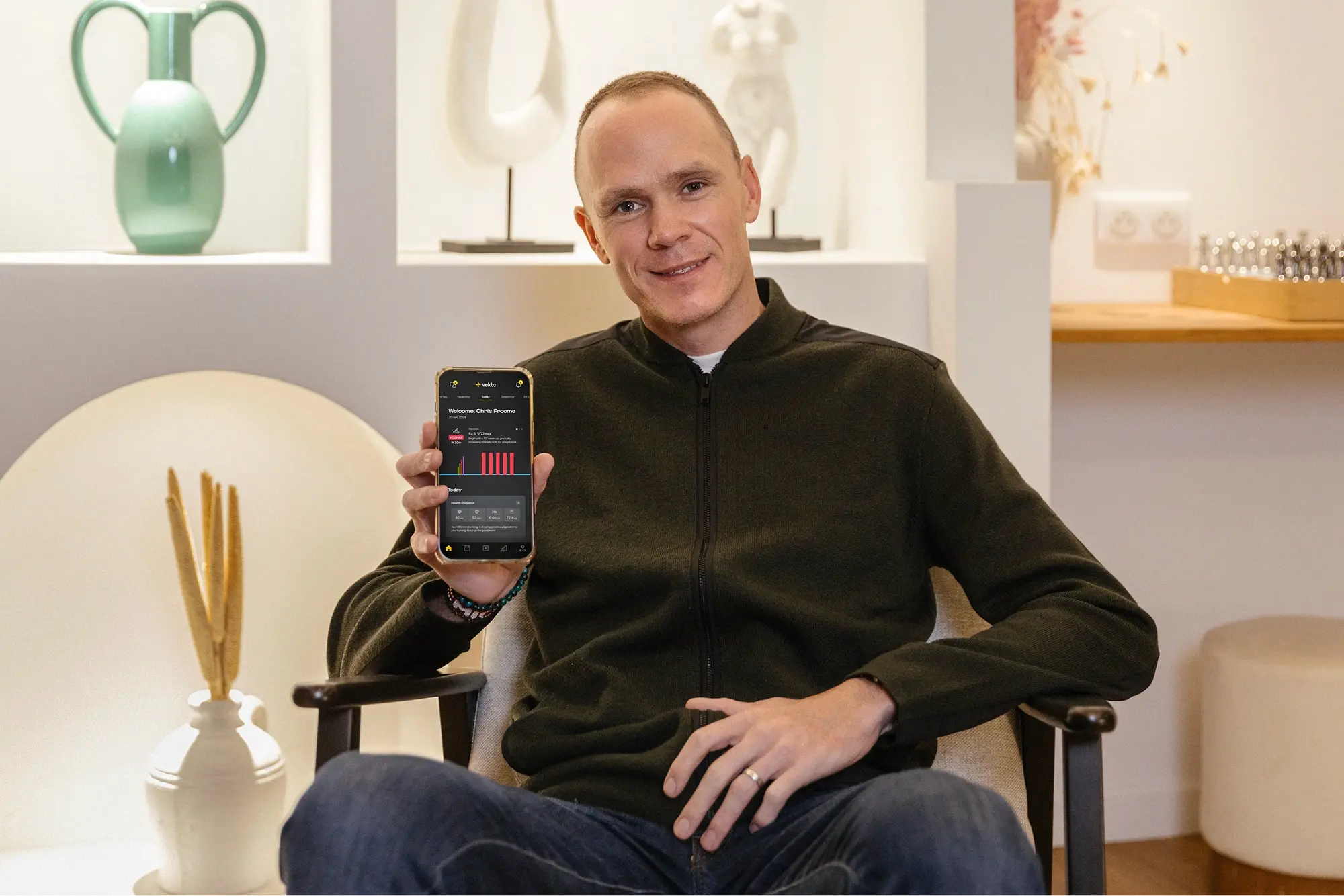 Chris Froome seated, holding a phone showing the Vekta mobile app open on his own athlete home screen