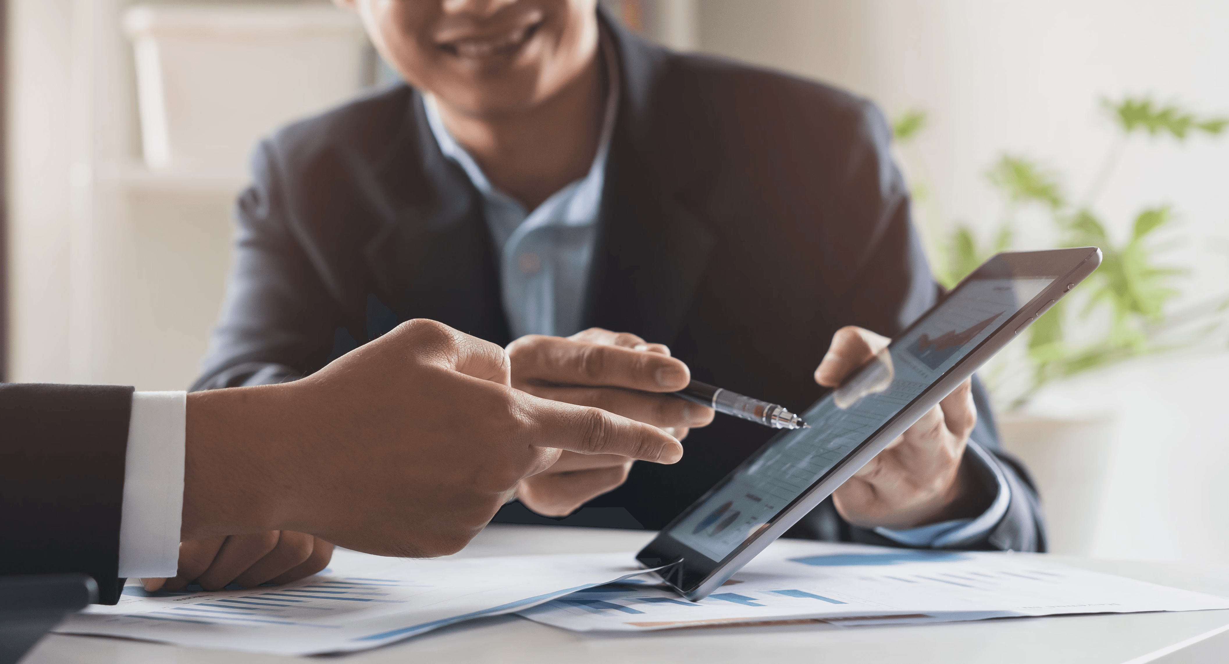 Zwei Geschäftsmänner im Anzug sitzen an einem Tisch und zeigen mit Stiften auf ein Tablet mit Diagrammen.