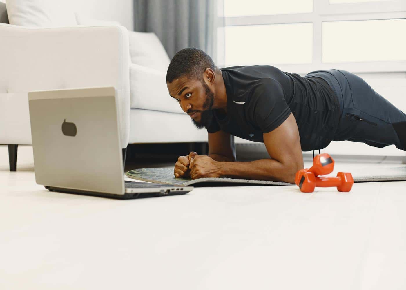 Man doing a plank on his forearms with weights next to him while staring at a laptop in front of him