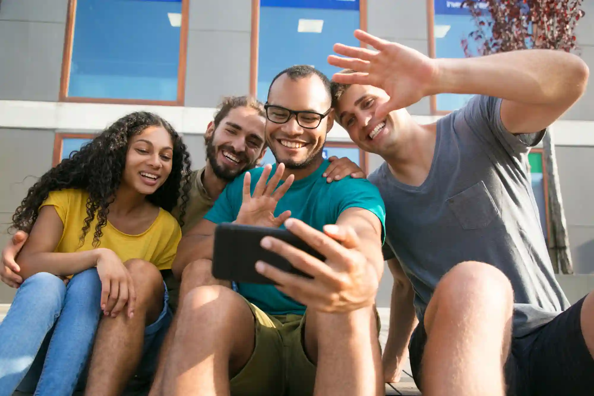 A diverse group of smiling friends wave while video chatting on a smartphone outdoors during a sunny day.