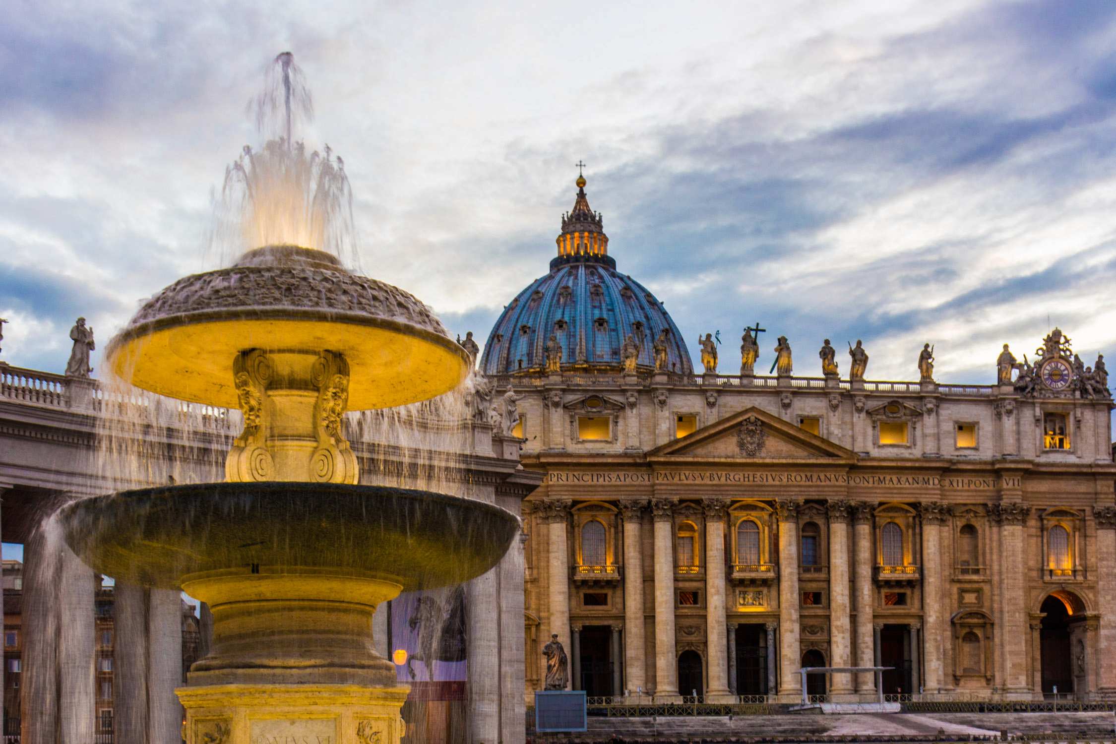A stone fountain with flowing water in the foreground and a large domed building under a cloudy blue sky.