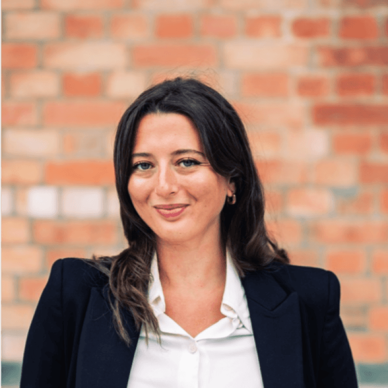 young brunette woman in a blazer and dress shirt in front of a brick wall