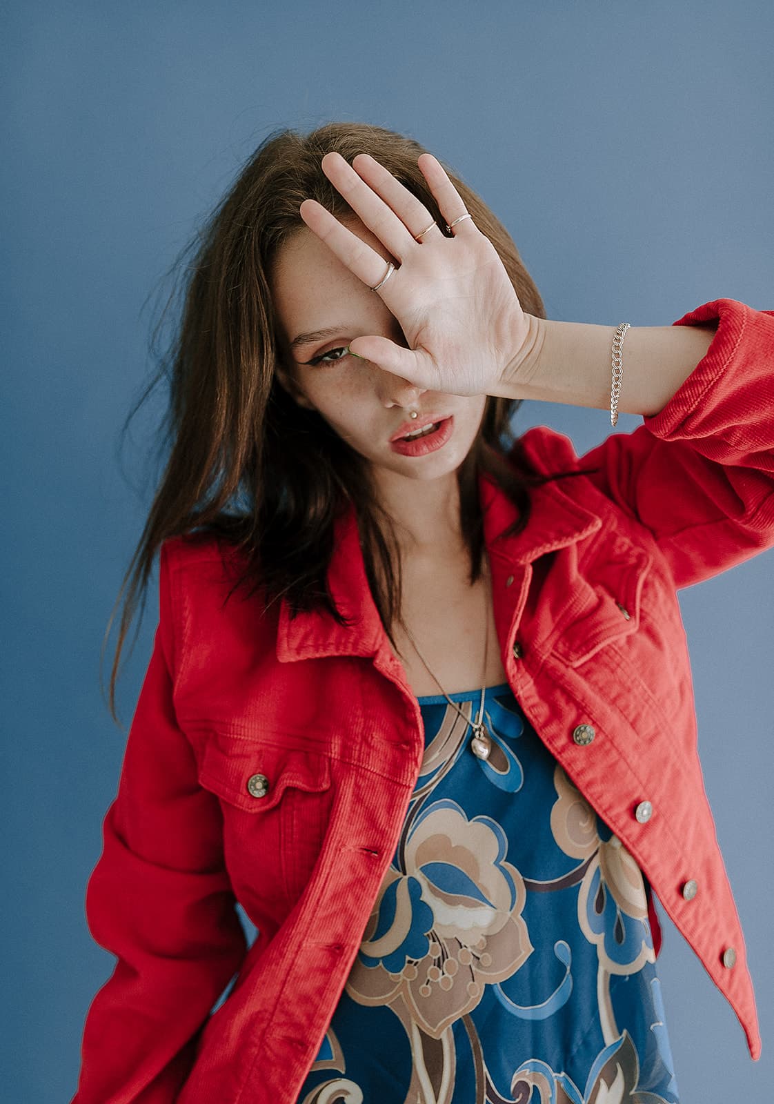 Model in red jacket poses with hand partially covering her face against a blue background at Revelator Studio’s natural light photography studio in Shreveport.