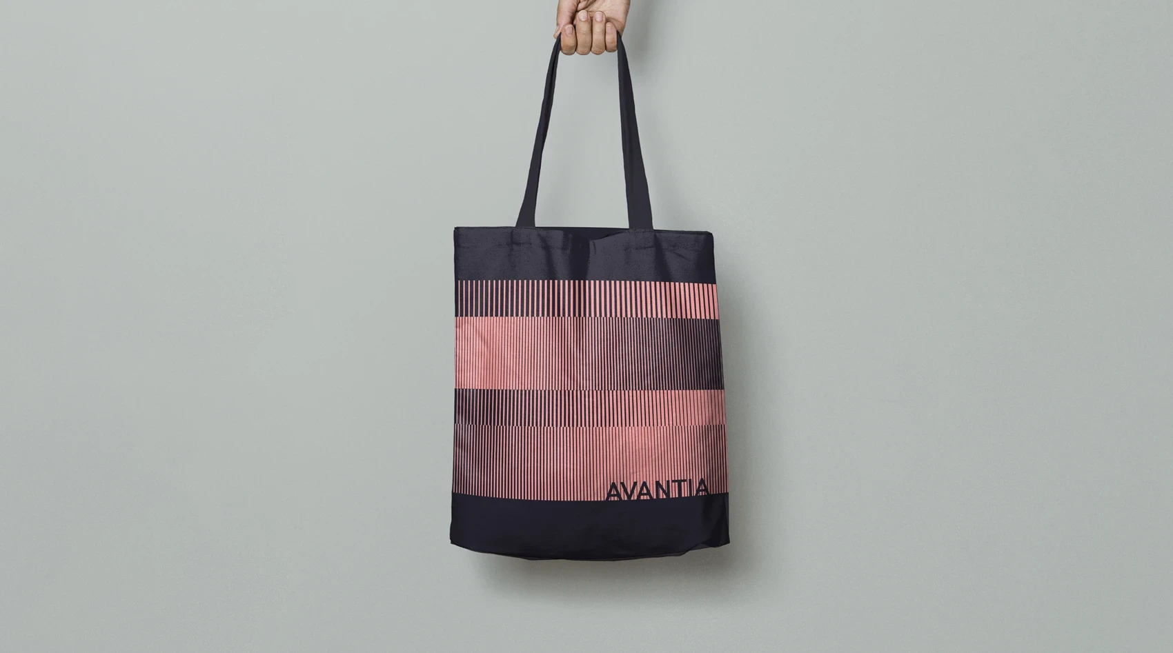 Black tote bag featuring horizontal bands of red and maroon vertical stripes. The ‘AVANTIA’ logo appears in black at the bottom right. A hand holds the bag up against a pale grey background.
