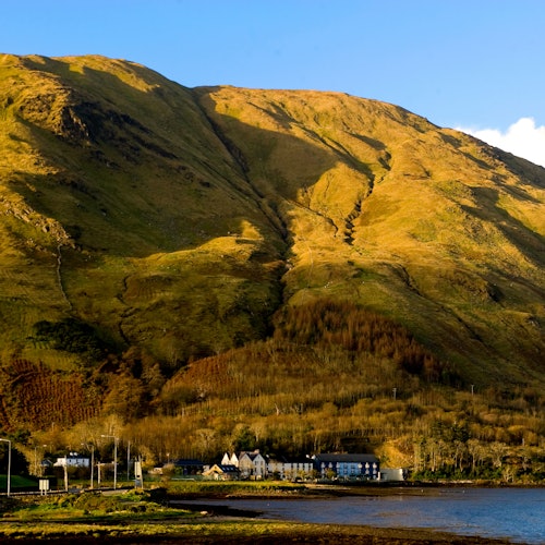 Село с няколко къщи в подножието на голяма, осветена от слънцето планина, със синьо небе отгоре. Воден басейн върху преден план.