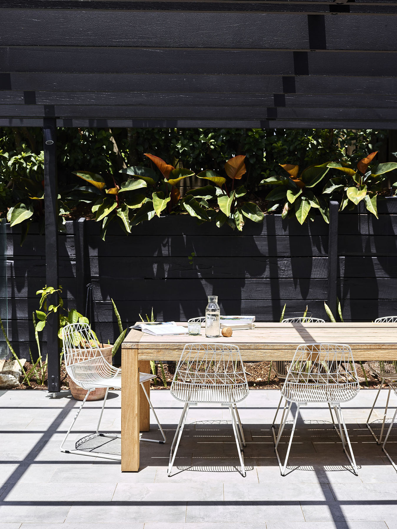 Outdoor dining area with timber table, white wire chairs, and garden backdrop.