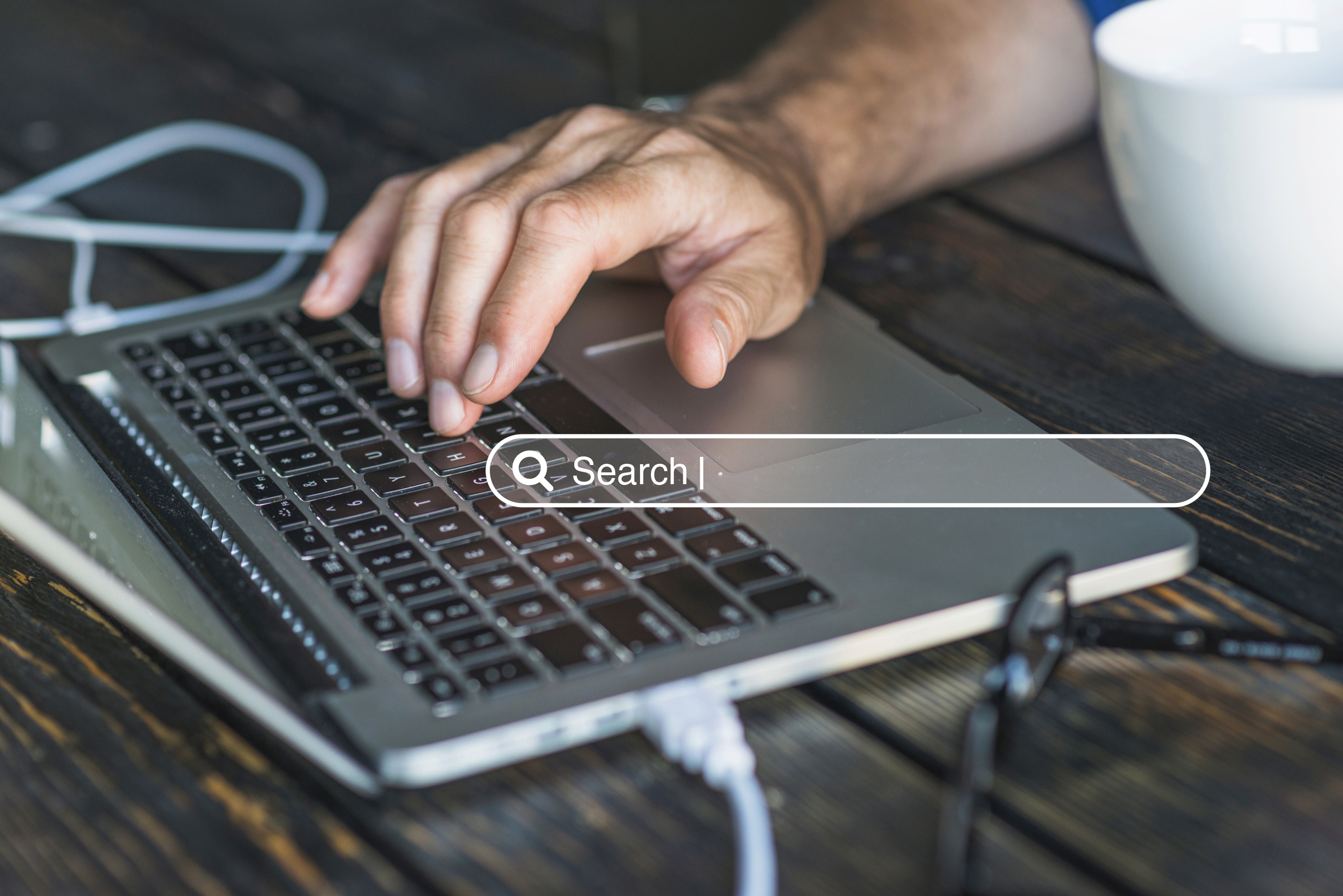 A close up of a person’s hand typing on a laptop keyboard on a wooden table, with a digital search bar graphic overlaid in the center of the image.