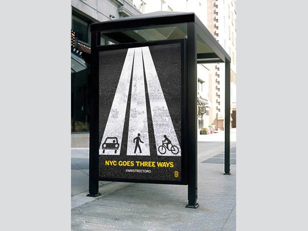 A black bus stop shelter stands on a city sidewalk. The shelter’s front panel displays a graphic of a crosswalk with three white stripes: a car on the left, a pedestrian in the middle, and a cyclist on the right. Below the graphic, the text reads “NYC GOES THREE WAYS” in yellow letters. Behind the shelter, buildings are visible, and the sidewalk is light gray.
