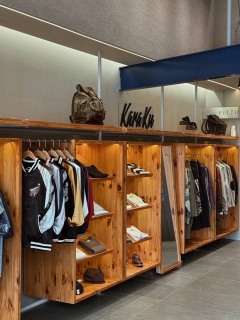 A retail display featuring wooden shelving showcasing various clothing items and accessories from Karaku Japanese Streetwear, including jackets, shirts, and bags.