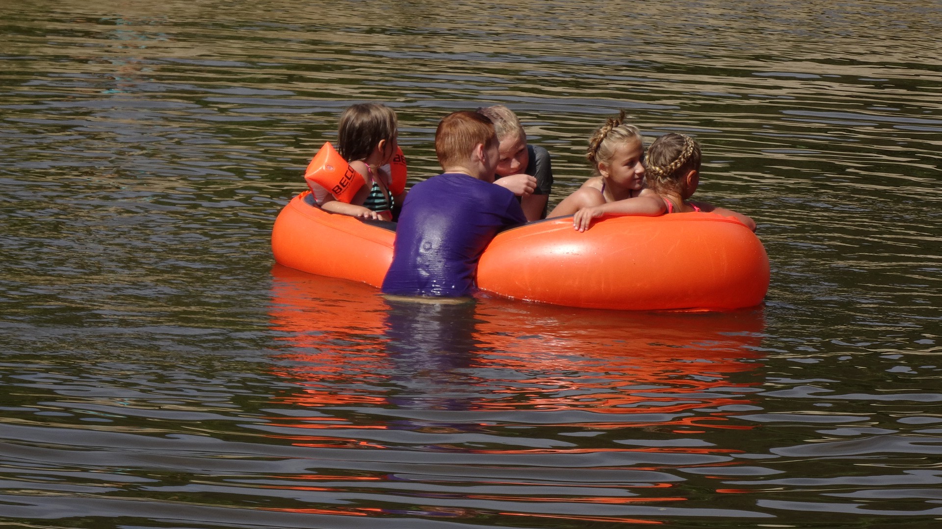 4 Kinder in einem roten Gummiboot auf dem See und ein Kind hängt im Wasser