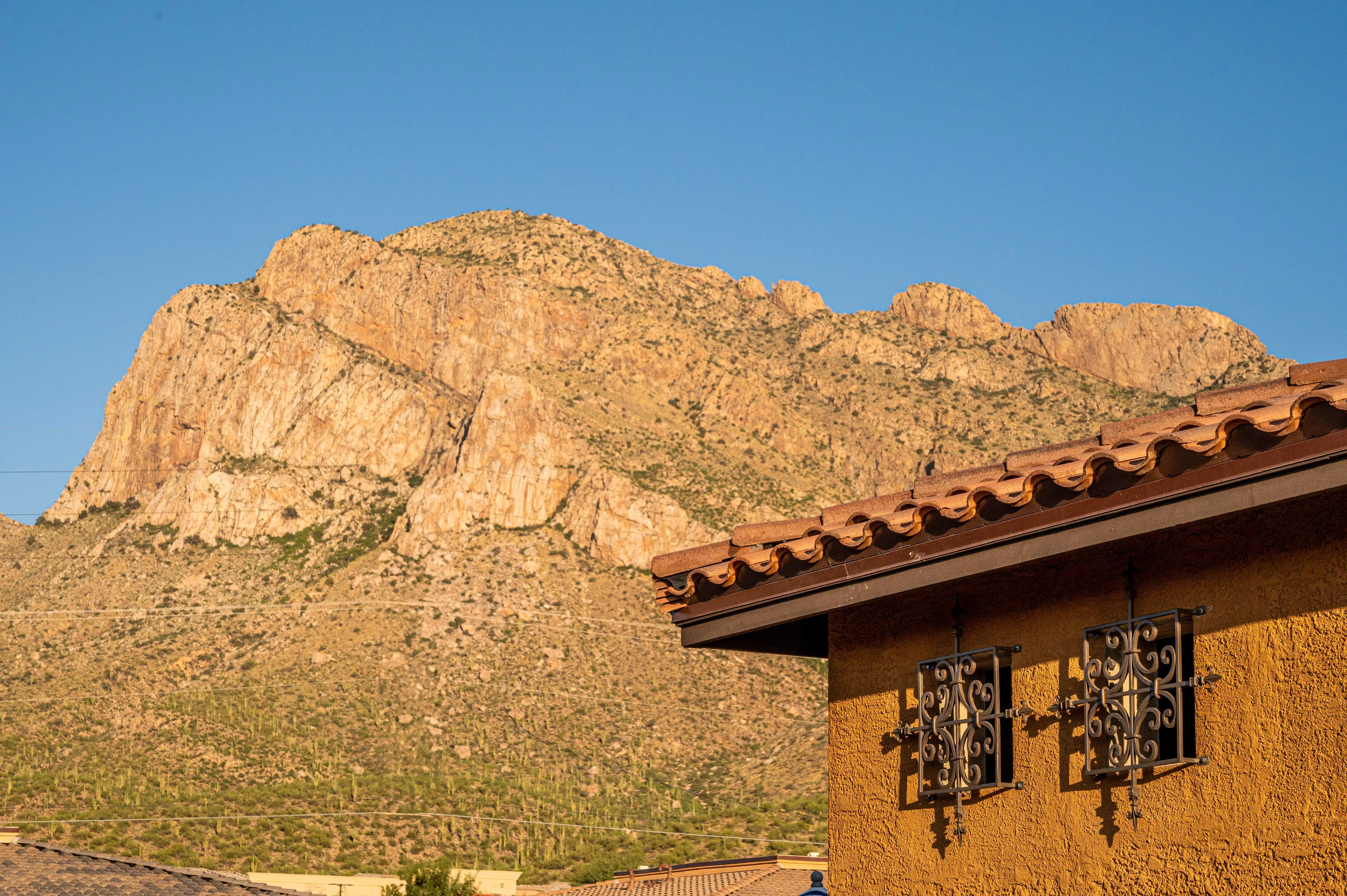 A desert mountain ridge under a clear blue sky with part of a stucco building featuring two wrought-iron window grills in the foreground