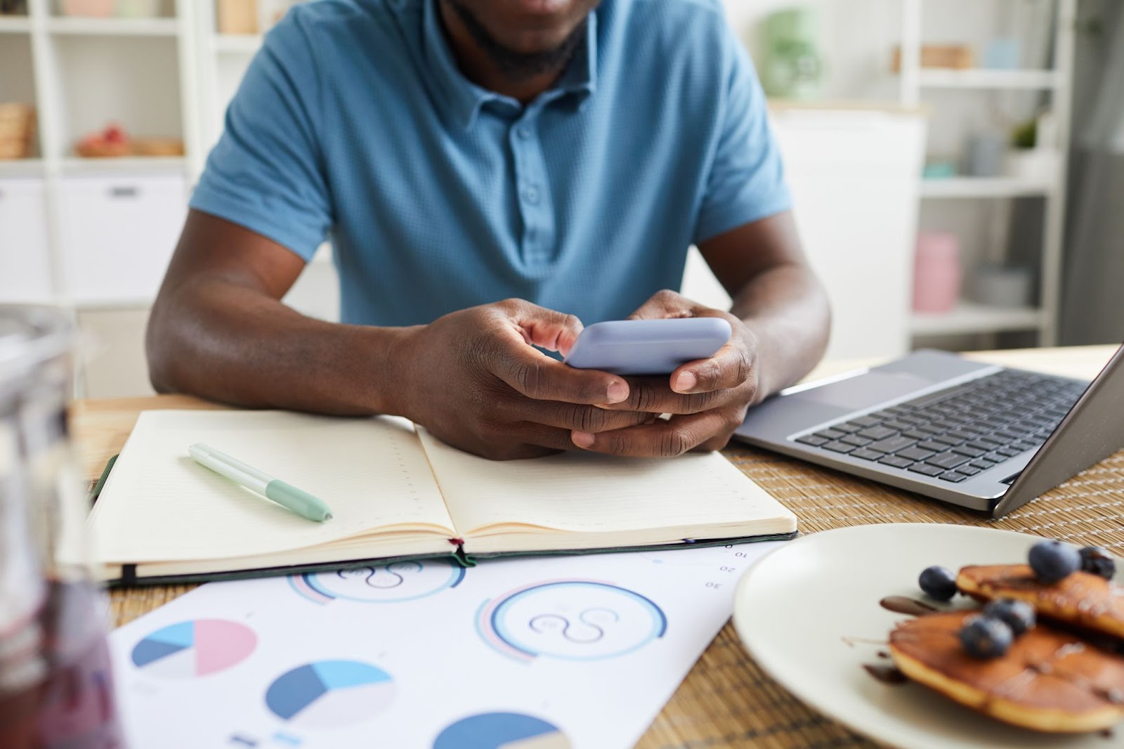 Man using a cell phone next to graphs, a notebook, and a laptop. 