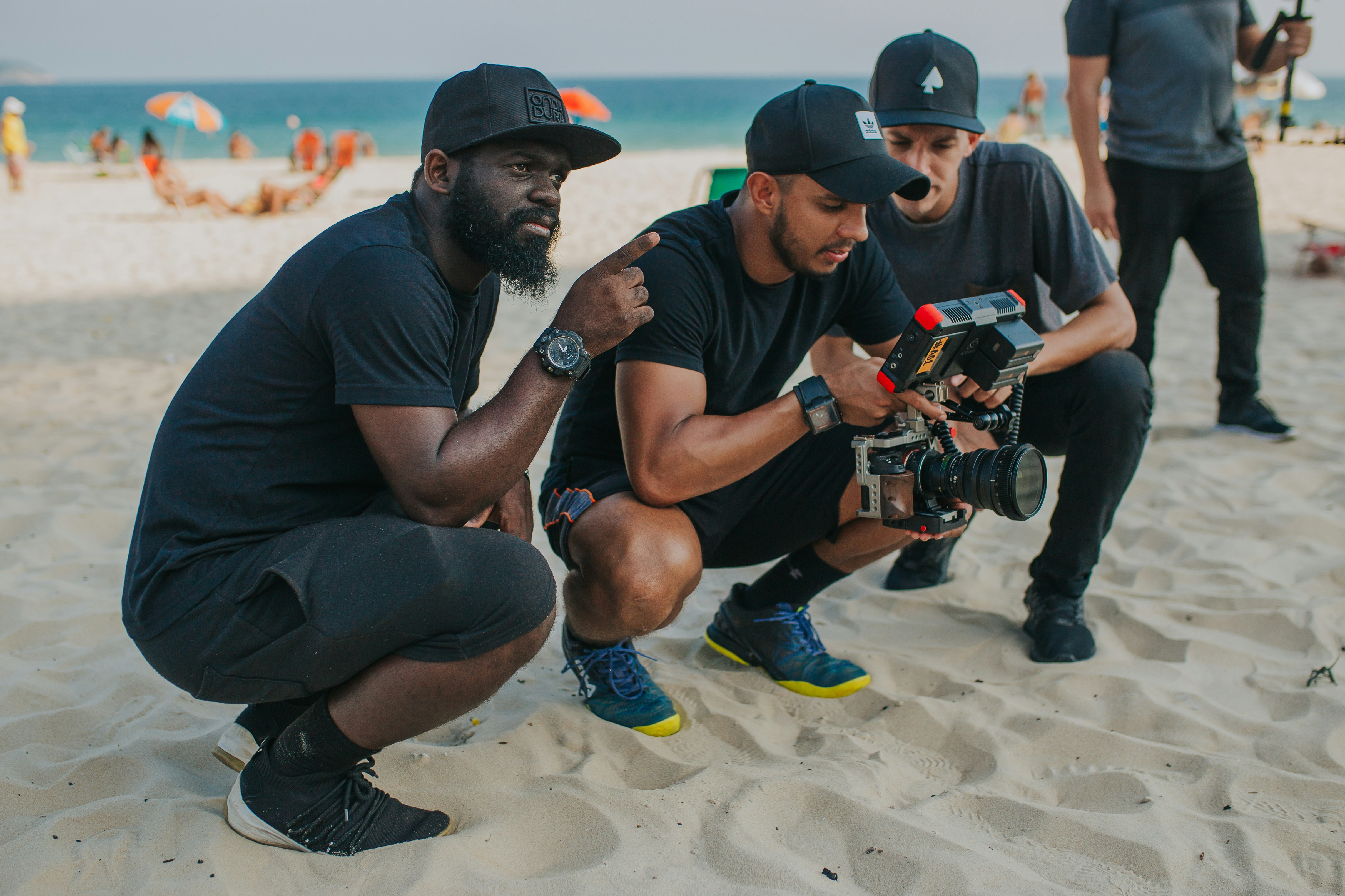 Three production crew members in black caps and athletic gear kneeling on a beach, reviewing footage on a professional cinema camera rig with an external monitor during a golden hour shoot.