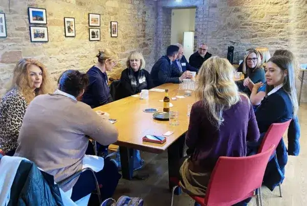 A group of people sit around a wooden table in a cozy, brick-walled room. They are engaged in lively conversation, creating a warm, collaborative atmosphere.