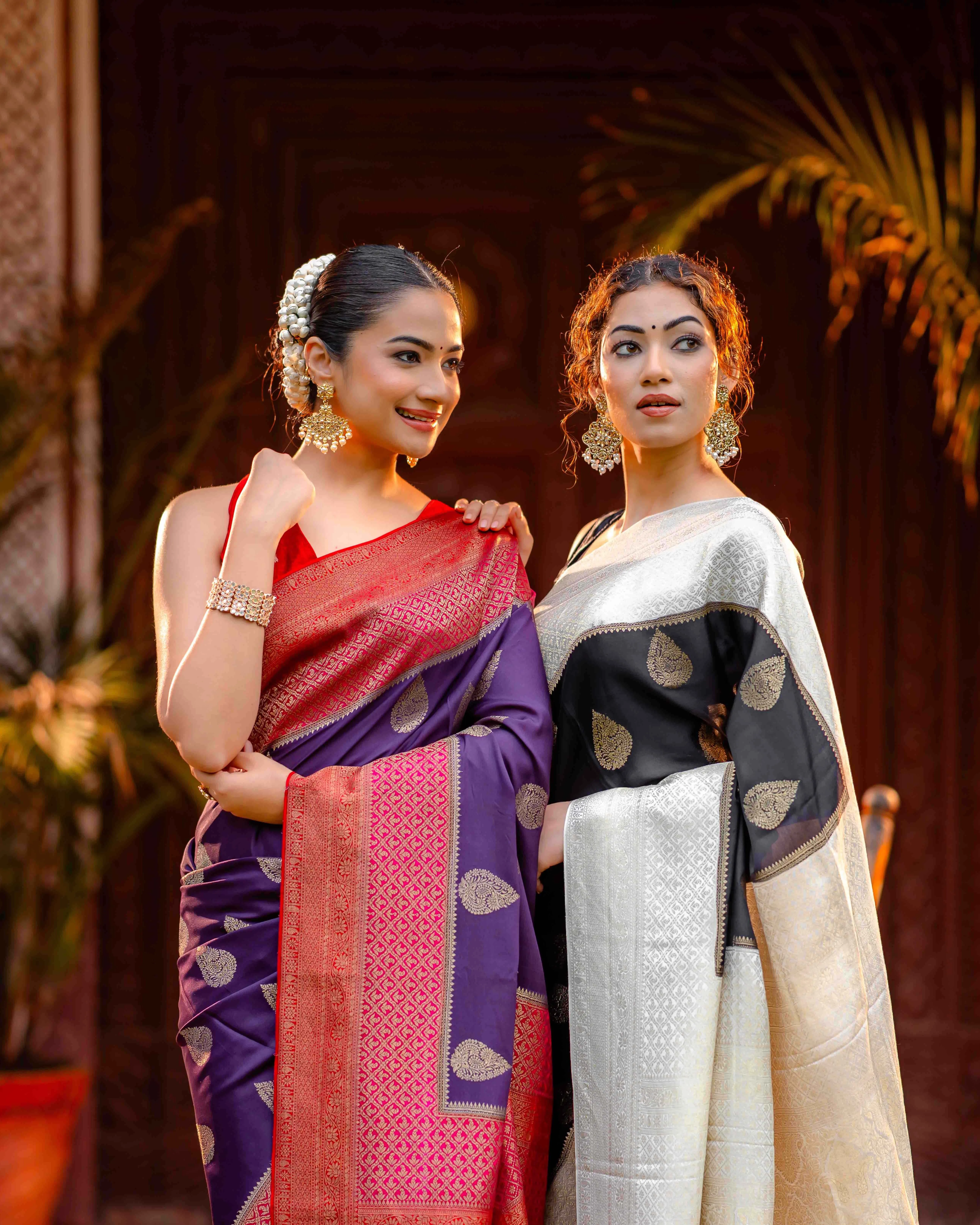 Two women standing together—one wearing a purple crepe silk saree with a pink-red contrast border and zari buttas, and the other wearing a black and white crepe saree—showcasing elegant handcrafted designs from That Saree Shop.