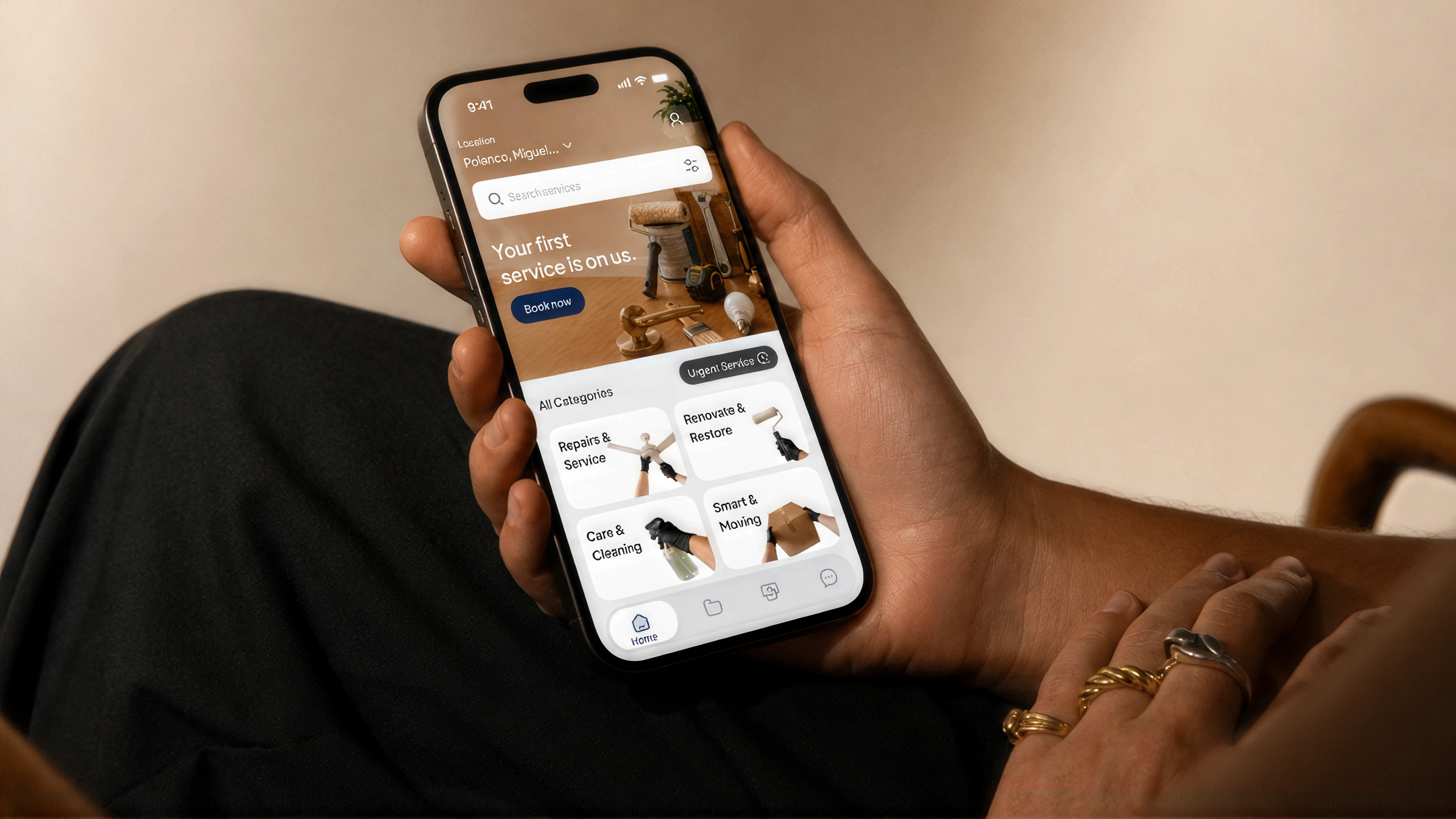 **Alt text:** A person holds a smartphone displaying a home-services app with a “Your first service is on us” promotion, search bar, and categories for repairs, renovation, cleaning, and moving services. The phone rests over the person’s lap in a warm indoor setting.