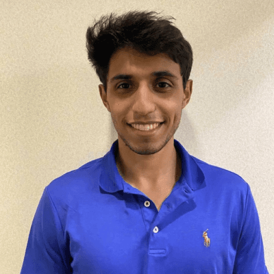Smiling Sebastian Capetta wearing a blue polo shirt stands against a plain light-colored wall, facing the camera in a simple portrait-style photo.