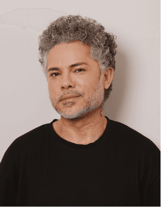 Retrato de um homem de cabelo grisalho e cacheado, com barba curta, vestindo camiseta preta em fundo neutro