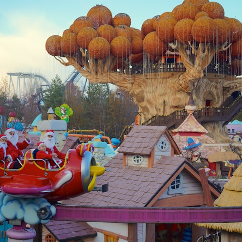 Der Weihnachtsmann und seine Helfer winken von einer Flugzeugfahrt in einem Vergnügungspark, der ein großes Baumhaus und skurrile Gebäude bietet.