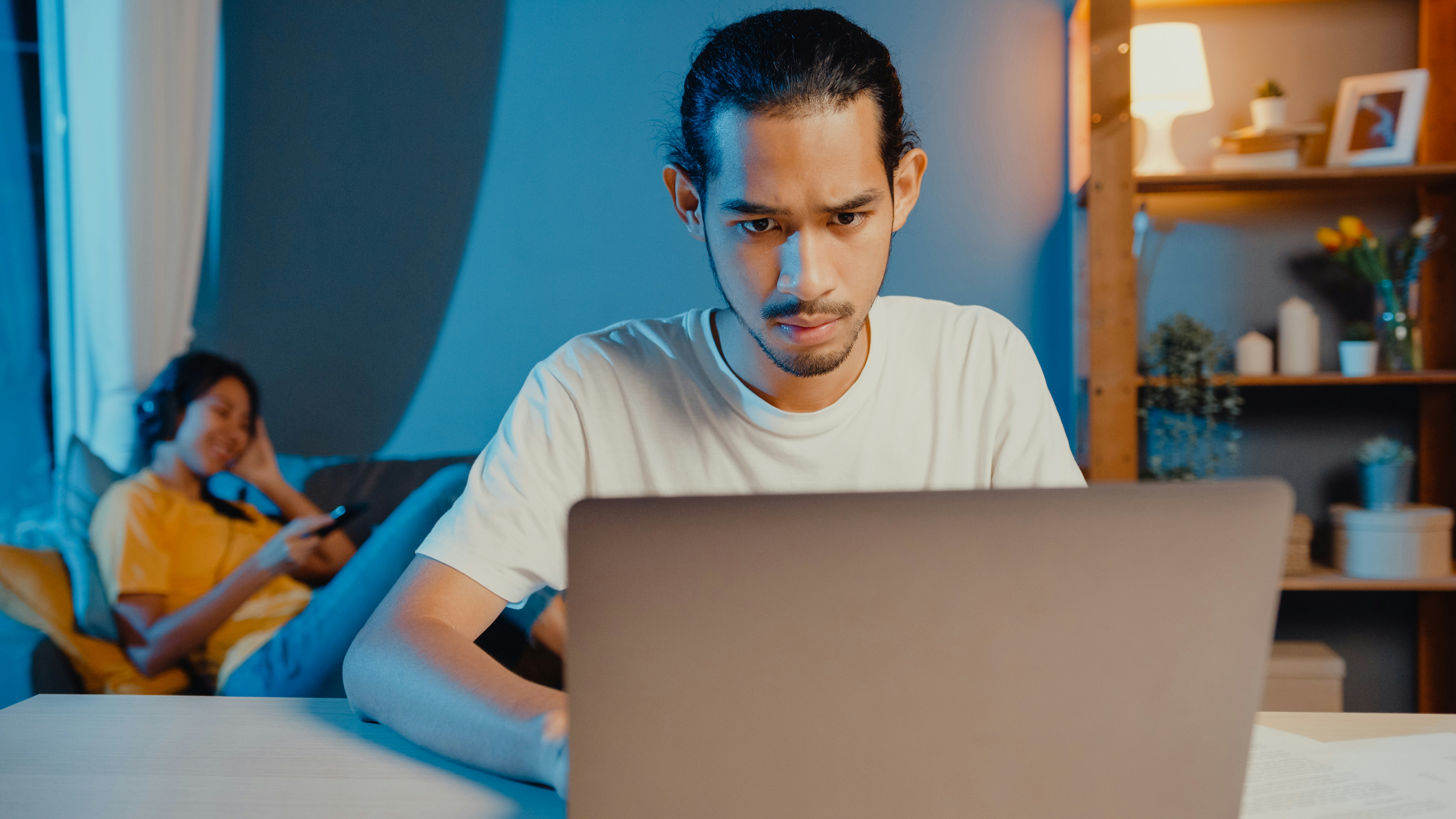 man using home computer with girl on couch in background wearing headphones