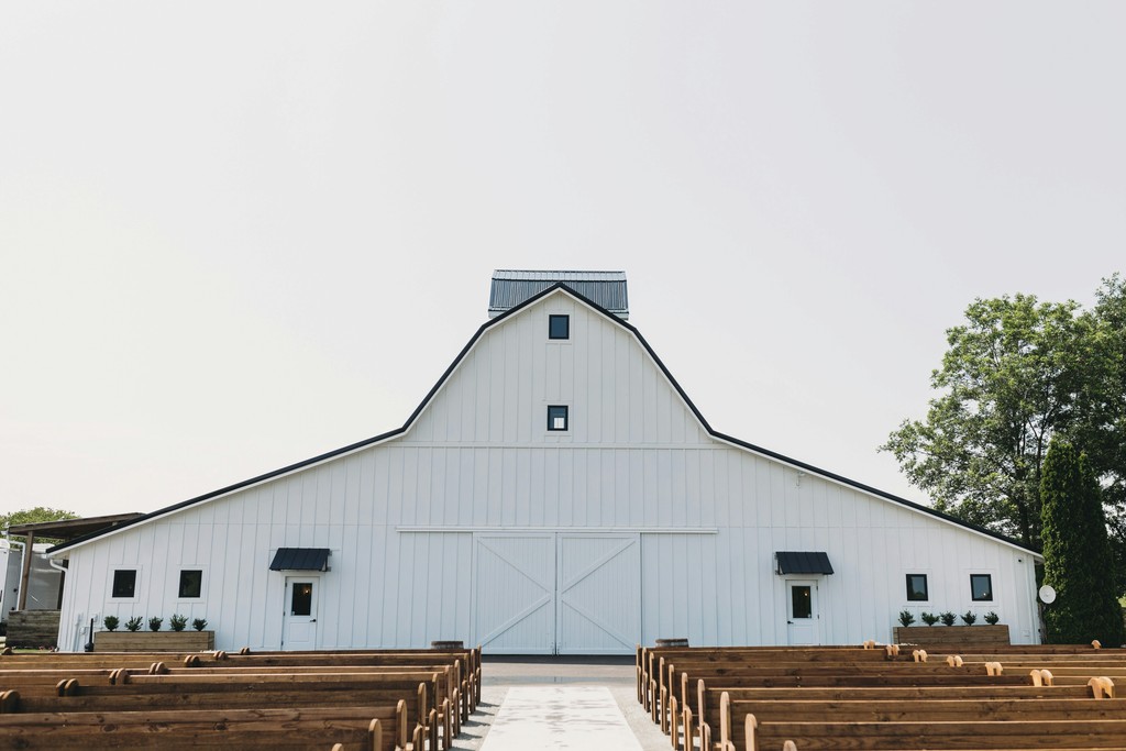 Outdoor ceremony aisle with rustic wooden pew benches leading to a white barn venue with crossbuck doors and black awnings
